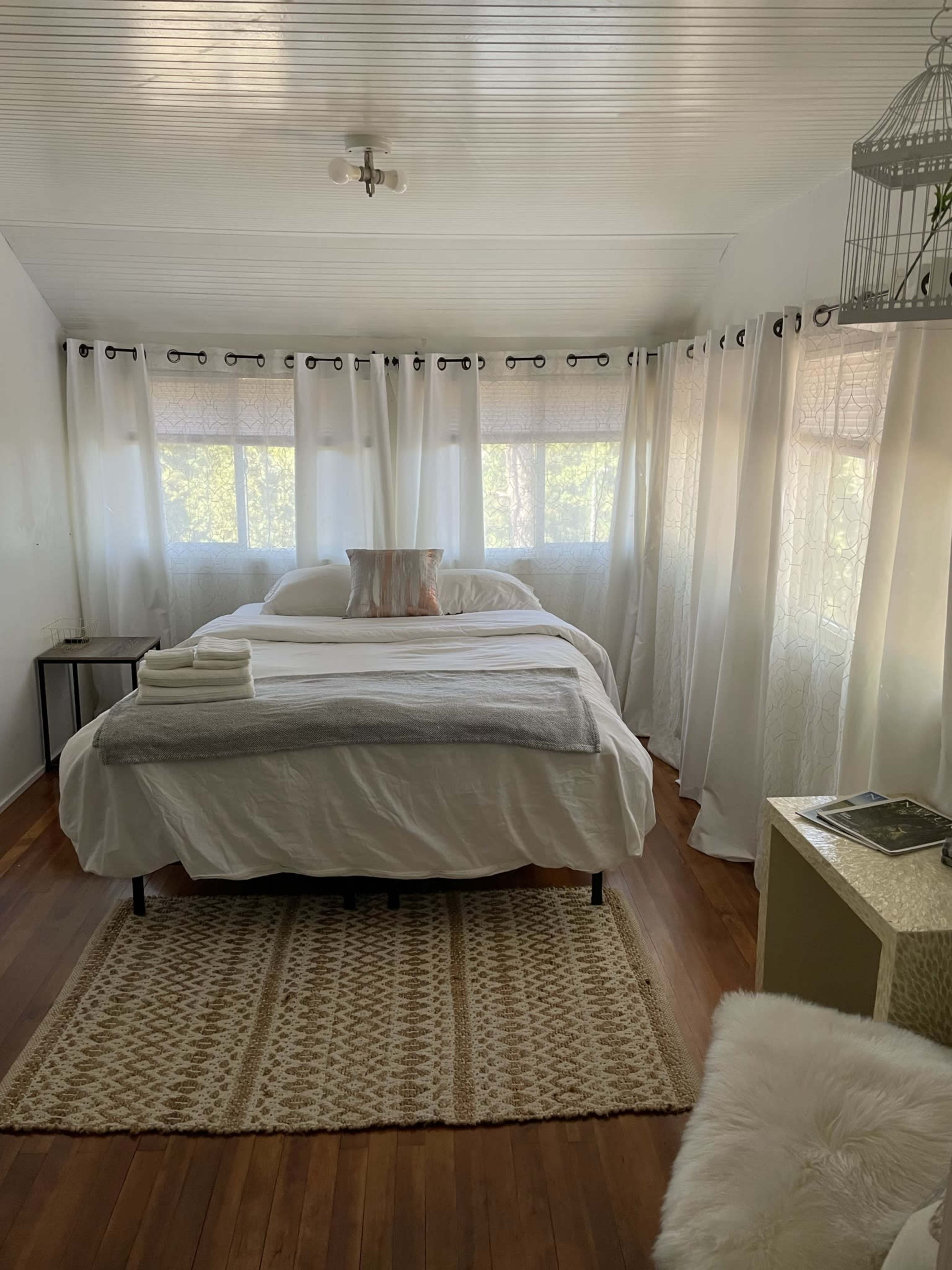 A bright, airy bedroom features two beds, large windows with sheer curtains, and a textured area rug on a hardwood floor.