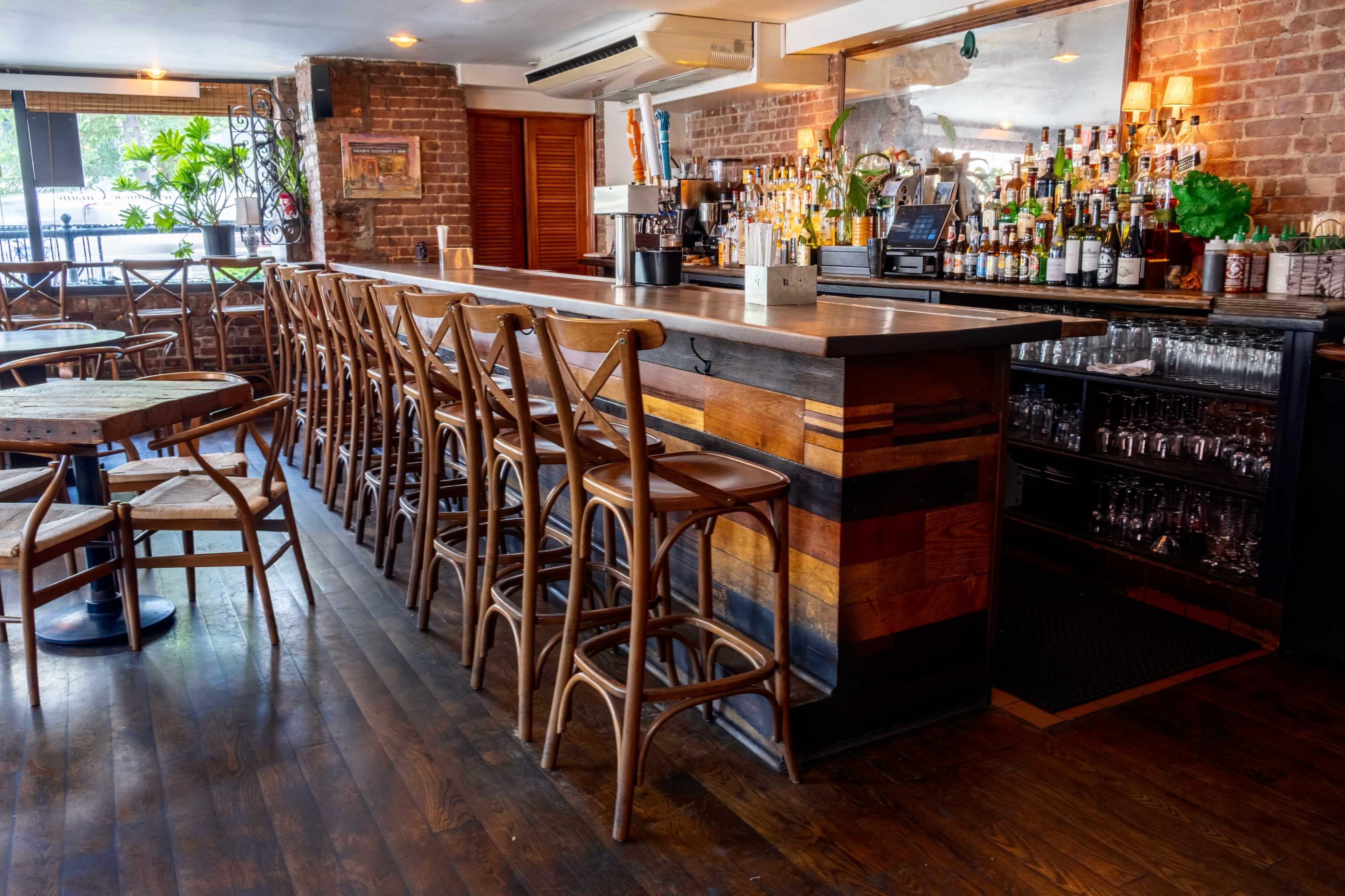 The image shows a bar area with a wooden counter, high stools arranged along it, and various bottles on shelves behind the bar against a brick wall.