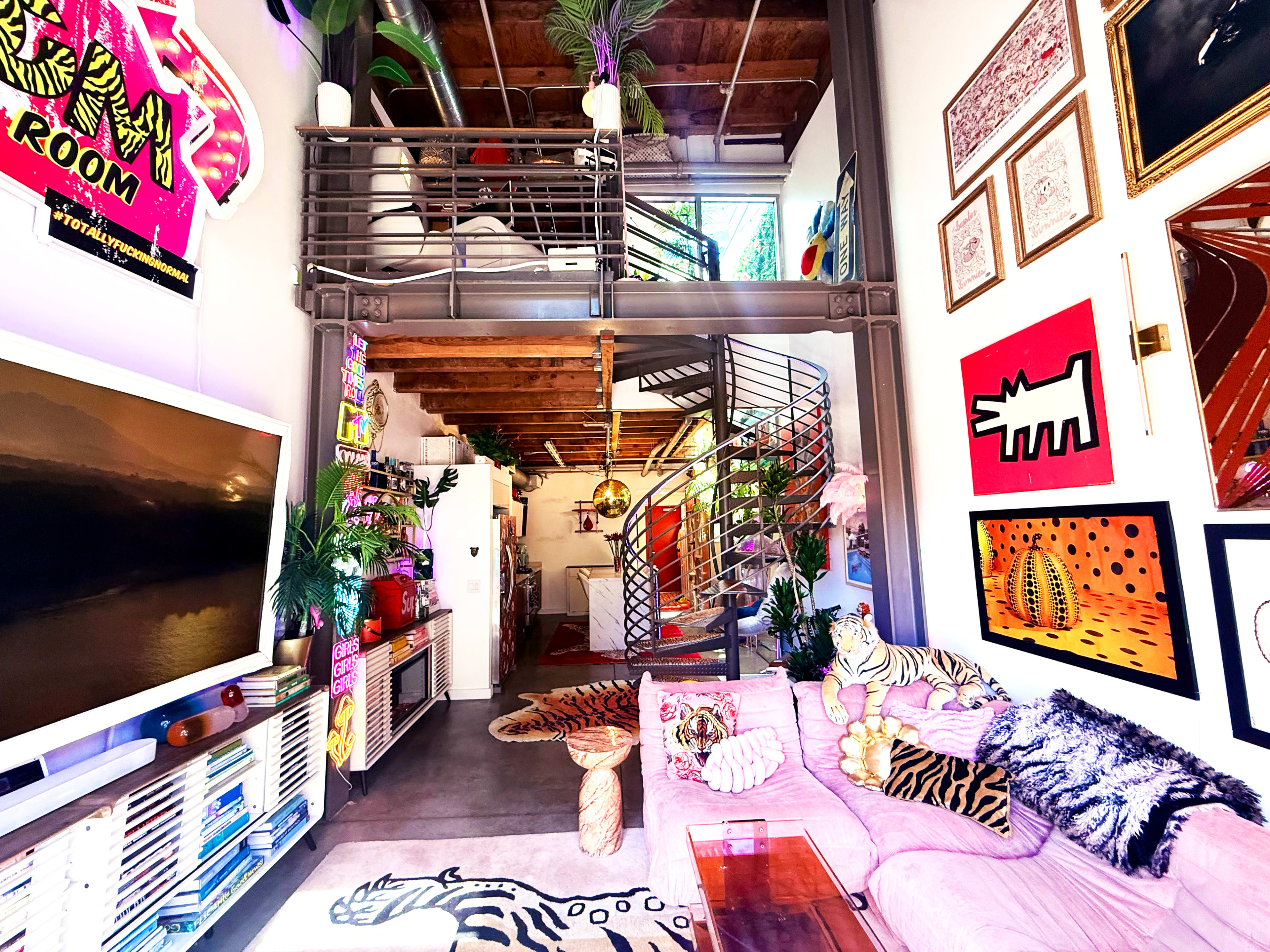 The image shows a vibrant, multi-level living space with eclectic decor, featuring a staircase, colorful artwork on the walls, and a mix of plant life and furniture.