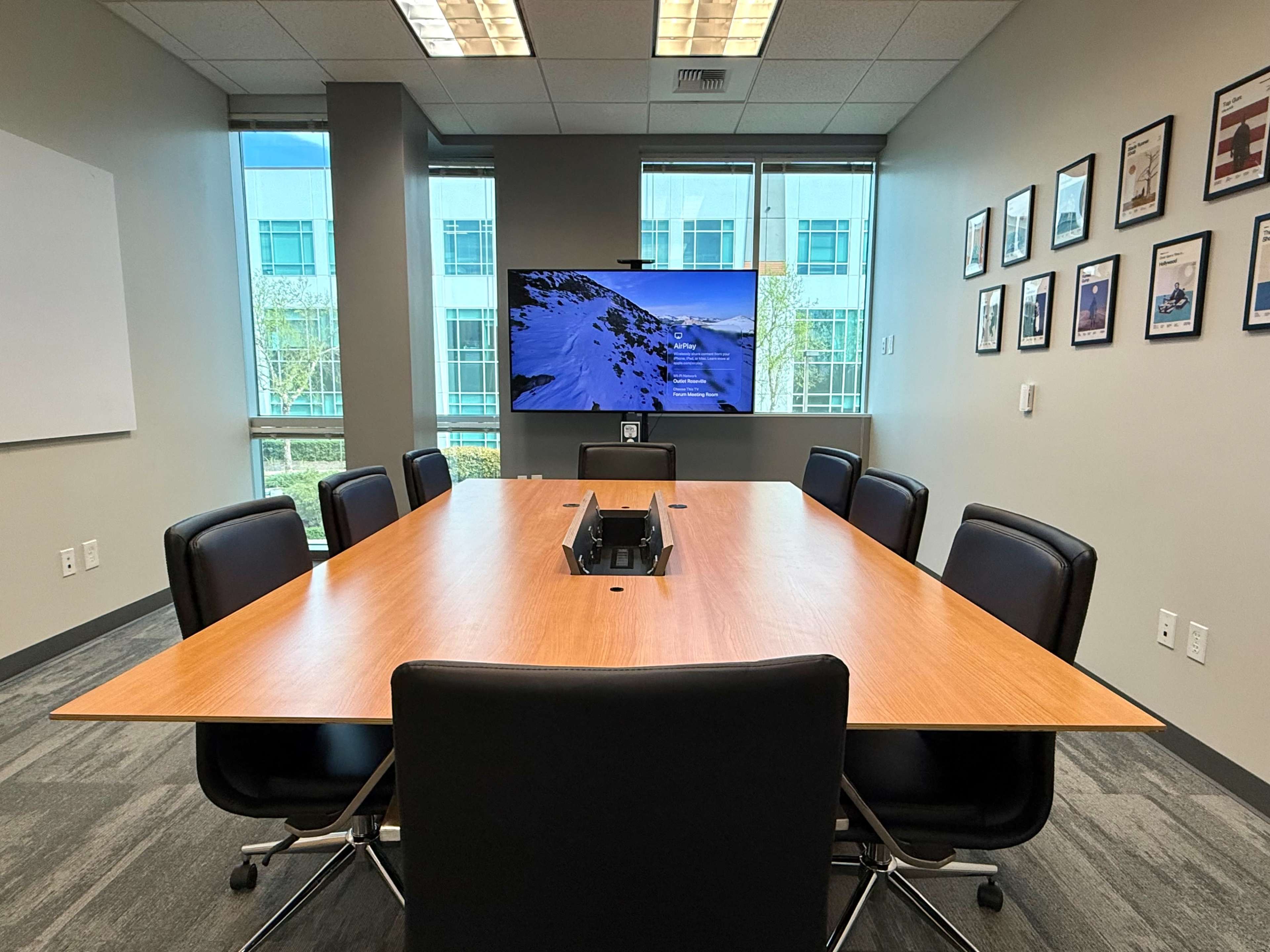 The image shows a modern conference room with a large wooden table, black chairs, and a wall-mounted television displaying a scenic image.