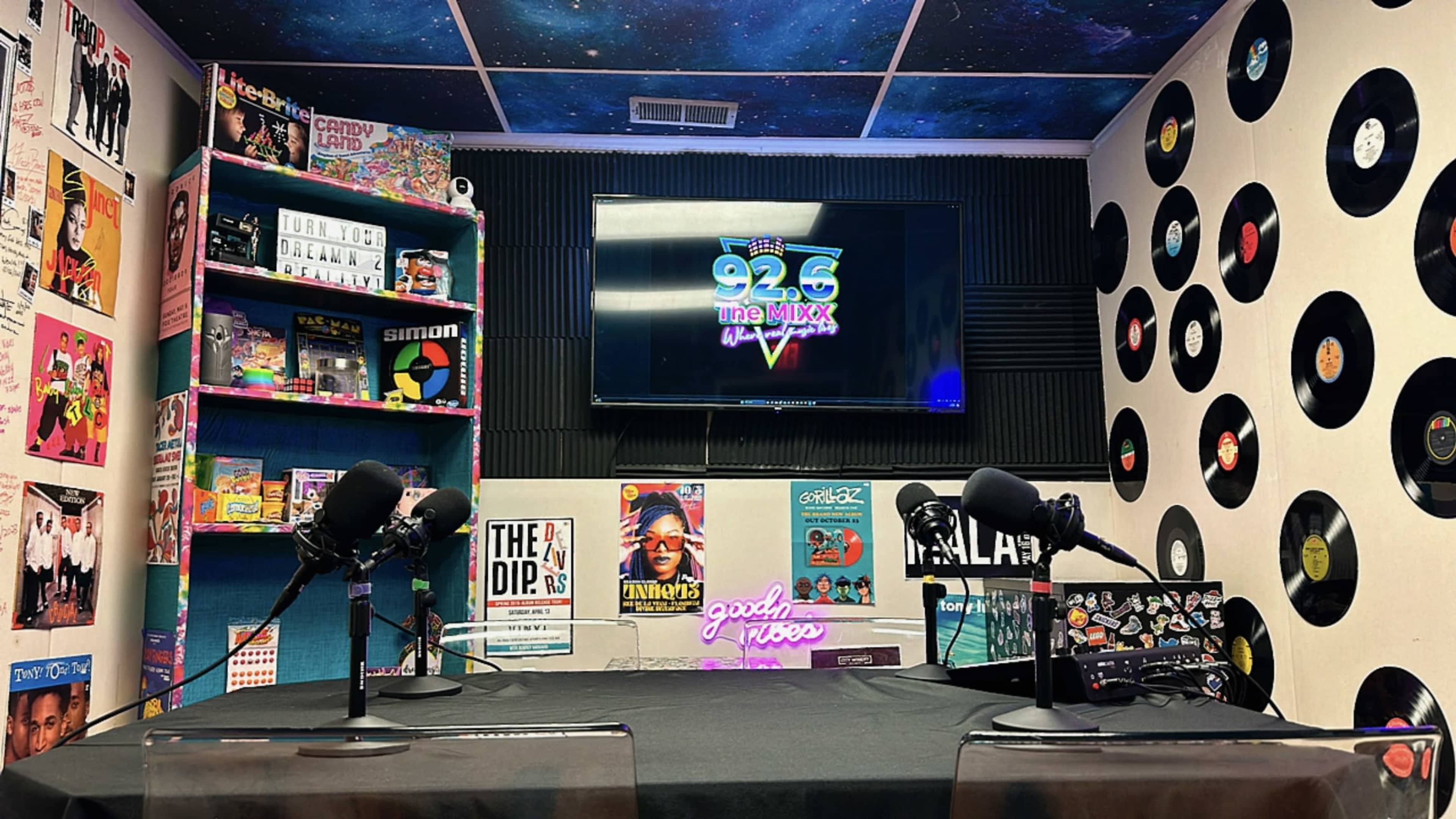 The image shows a radio studio with a dark table, two microphones, colorful record decorations on the walls, and a television displaying the station's logo.