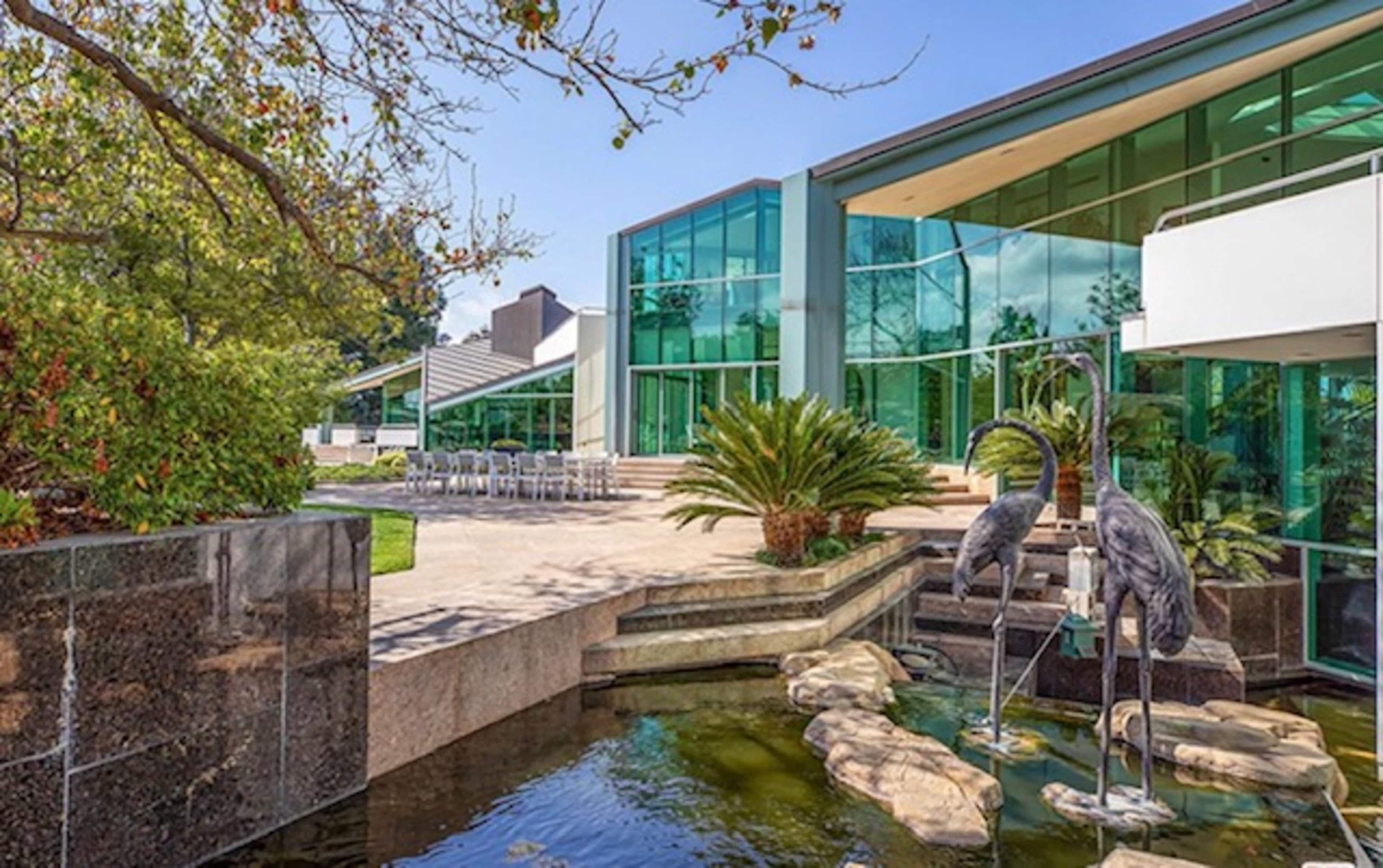 A modern house with large glass windows, a stone pond featuring two crane sculptures, and a patio area surrounded by greenery.