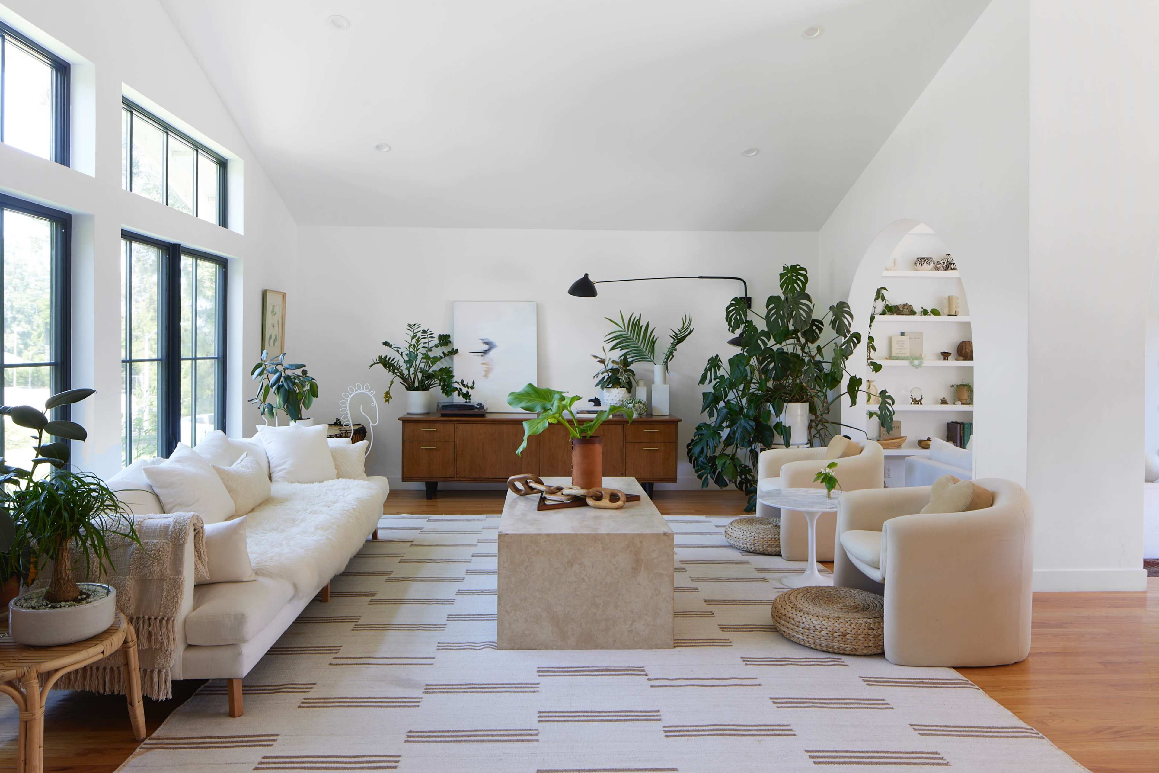 A modern living room featuring a light sectional sofa, a large stone coffee table, and various plants arranged around a wooden sideboard.