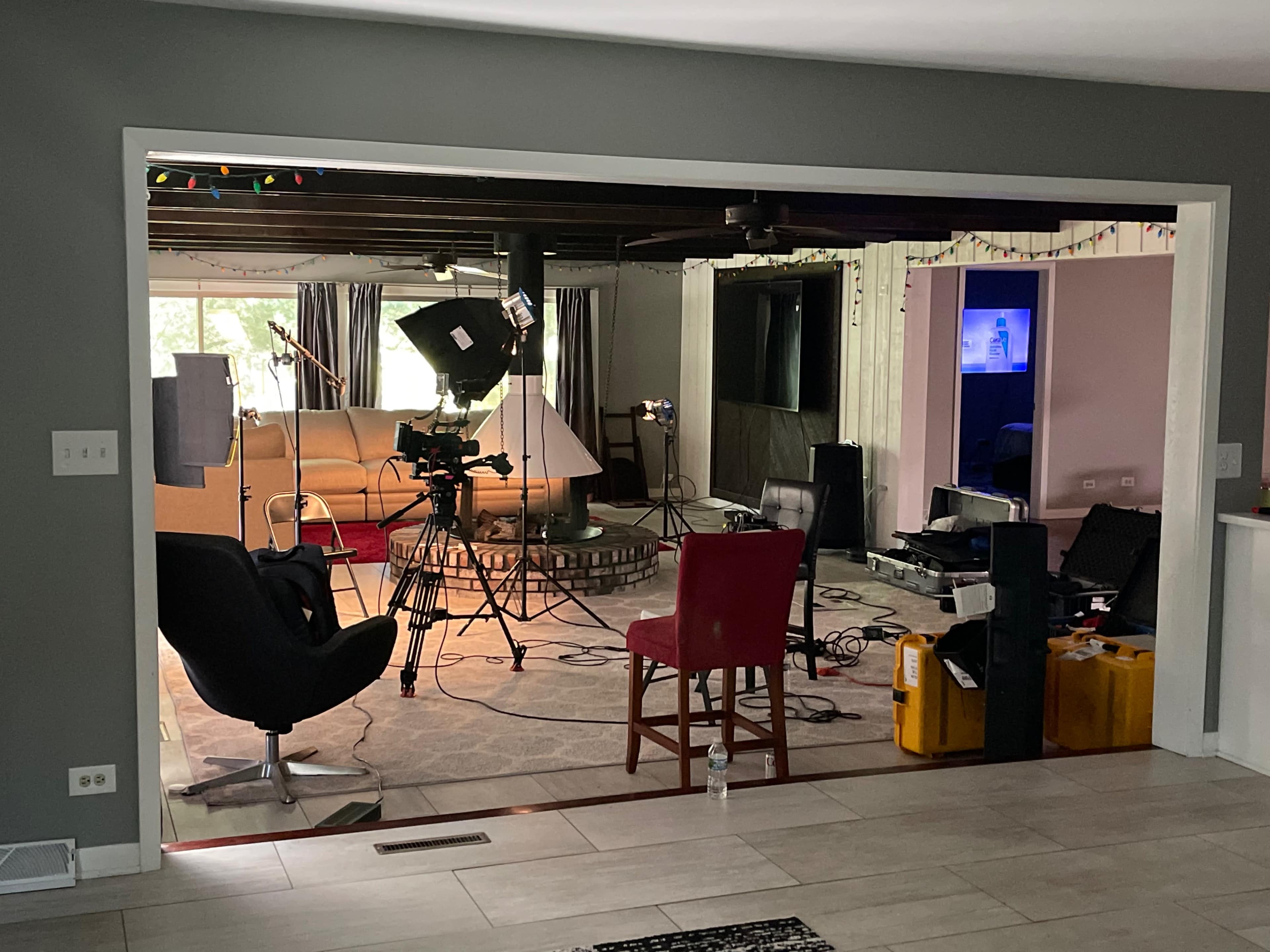 A film crew sets up equipment in a living room with a visible sofa, lighting, and cameras arranged for a shoot.