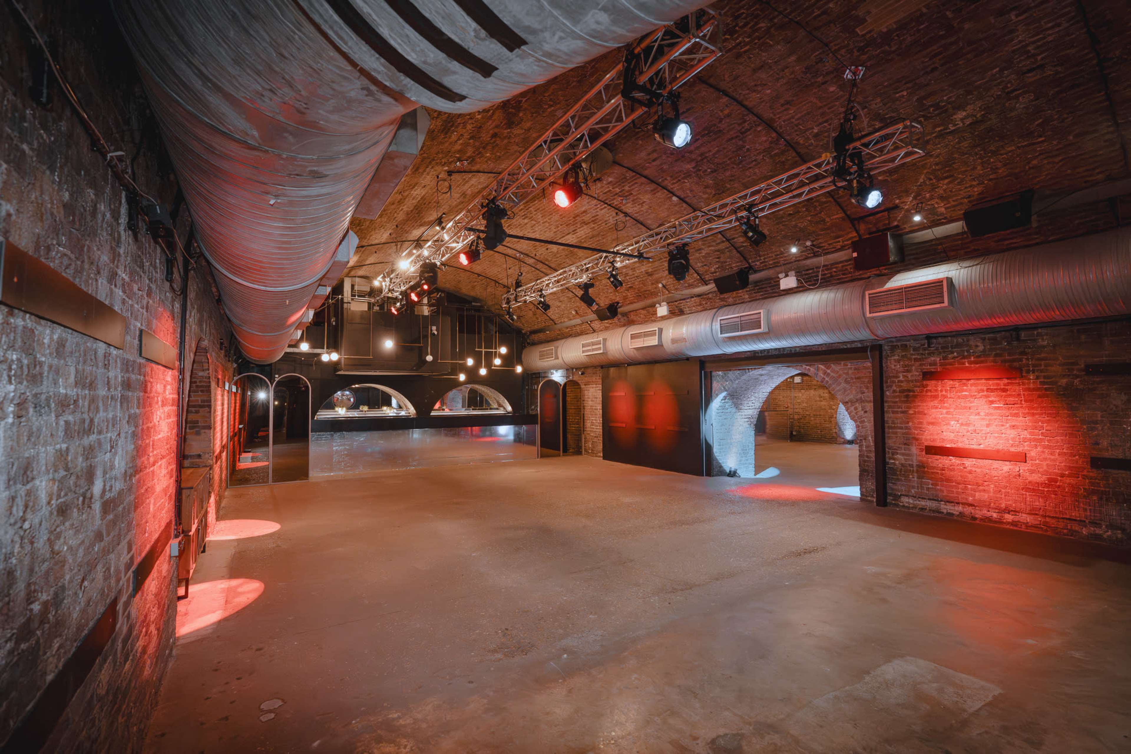 The image shows an empty event space with high ceilings, exposed brick walls, and a series of spotlights and colorful lights illuminating the area.