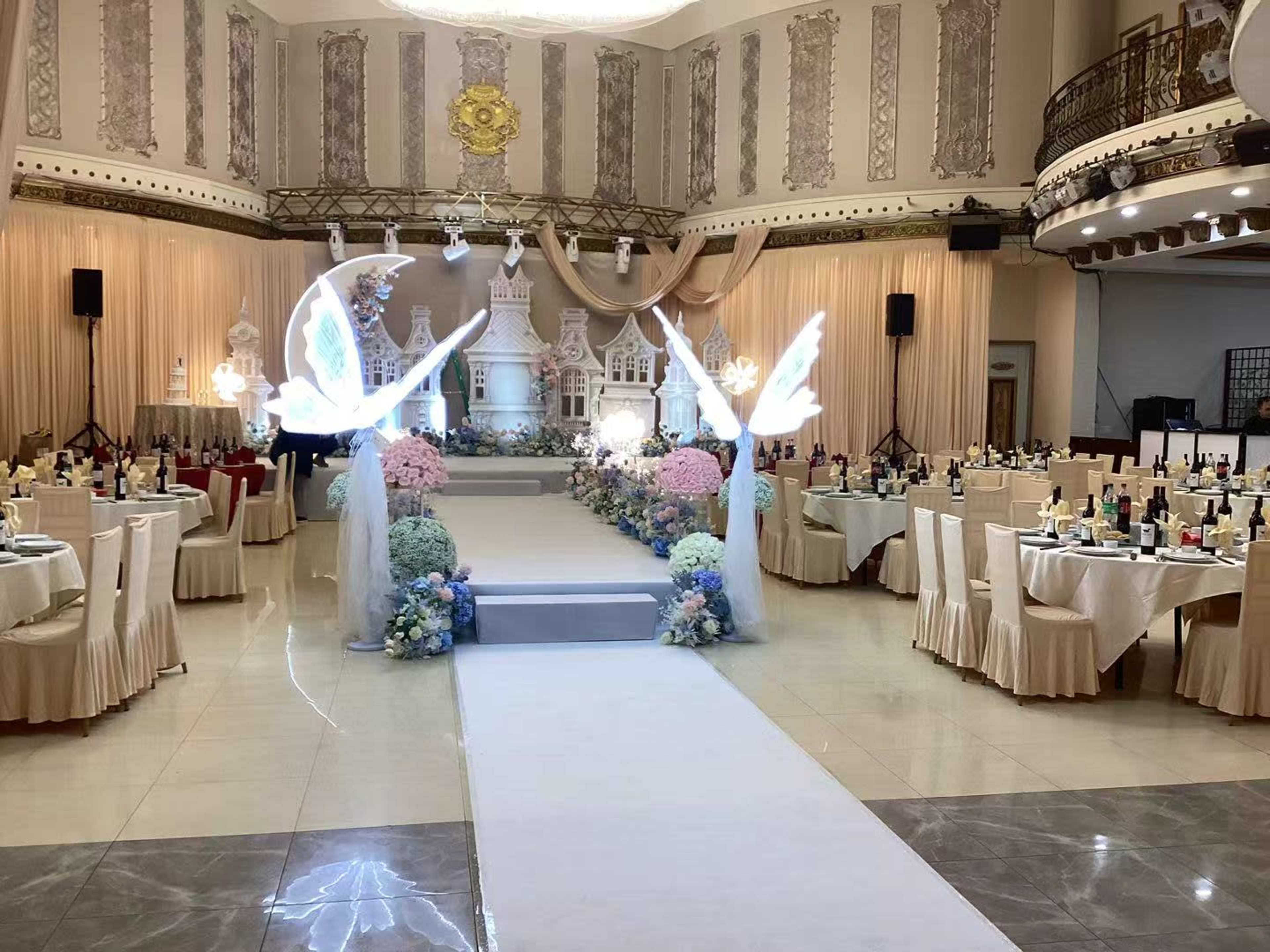 The image depicts an elegantly set banquet hall with tables arranged around a central aisle, adorned with floral decorations and illuminated displays.