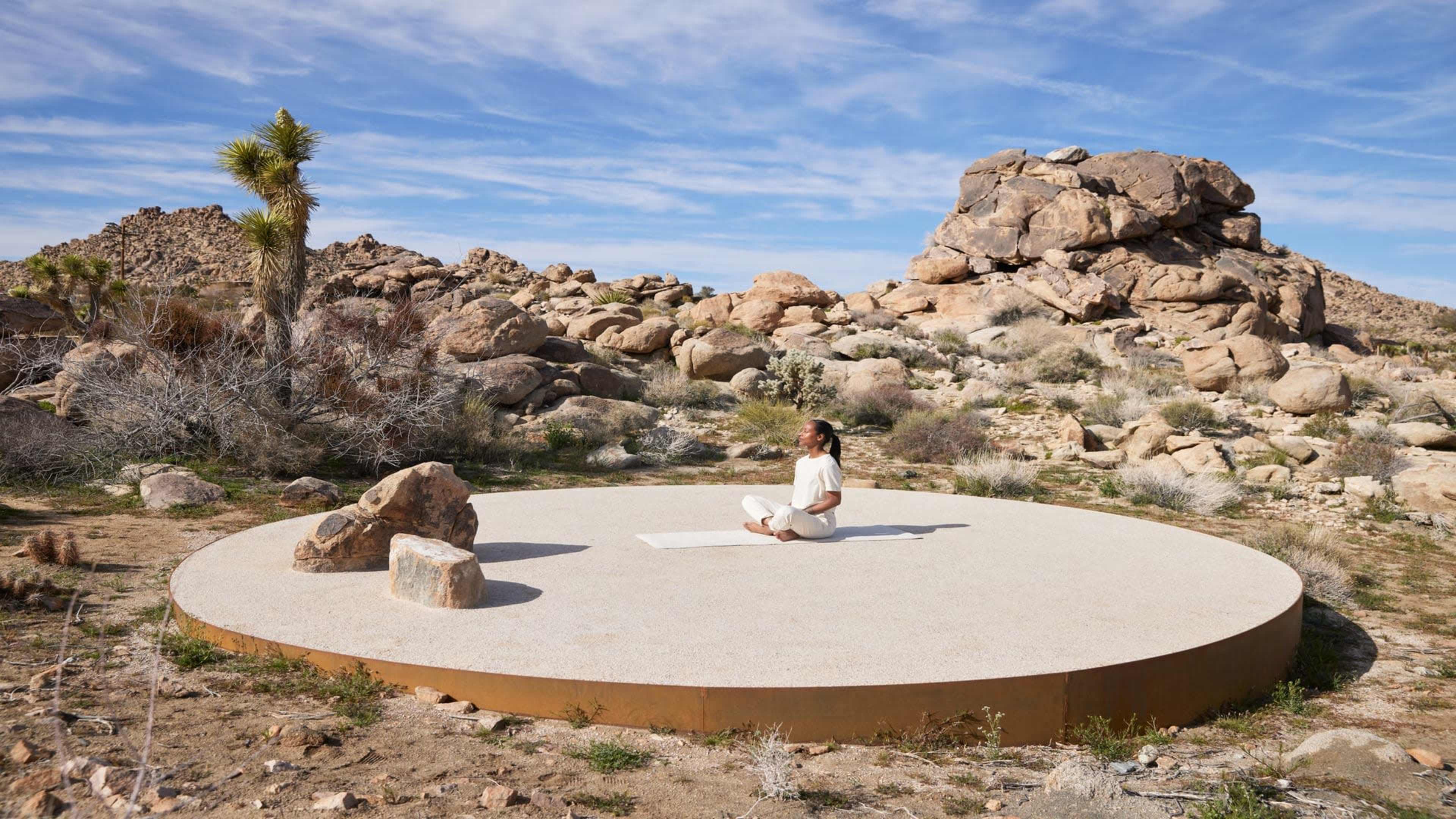 A person performs yoga on a circular platform surrounded by rocks and desert vegetation.