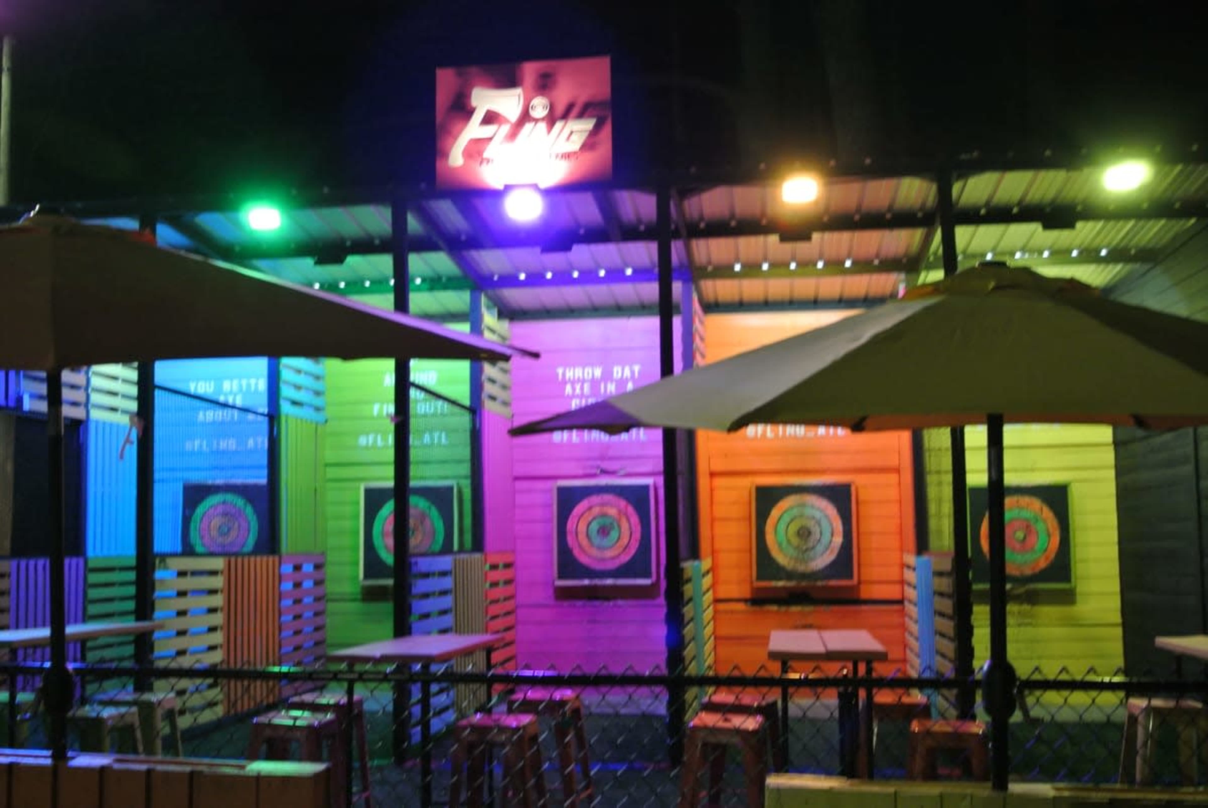 The image shows a brightly lit outdoor throwing range with colorful walls, dartboards, and seating arrangements under umbrellas.