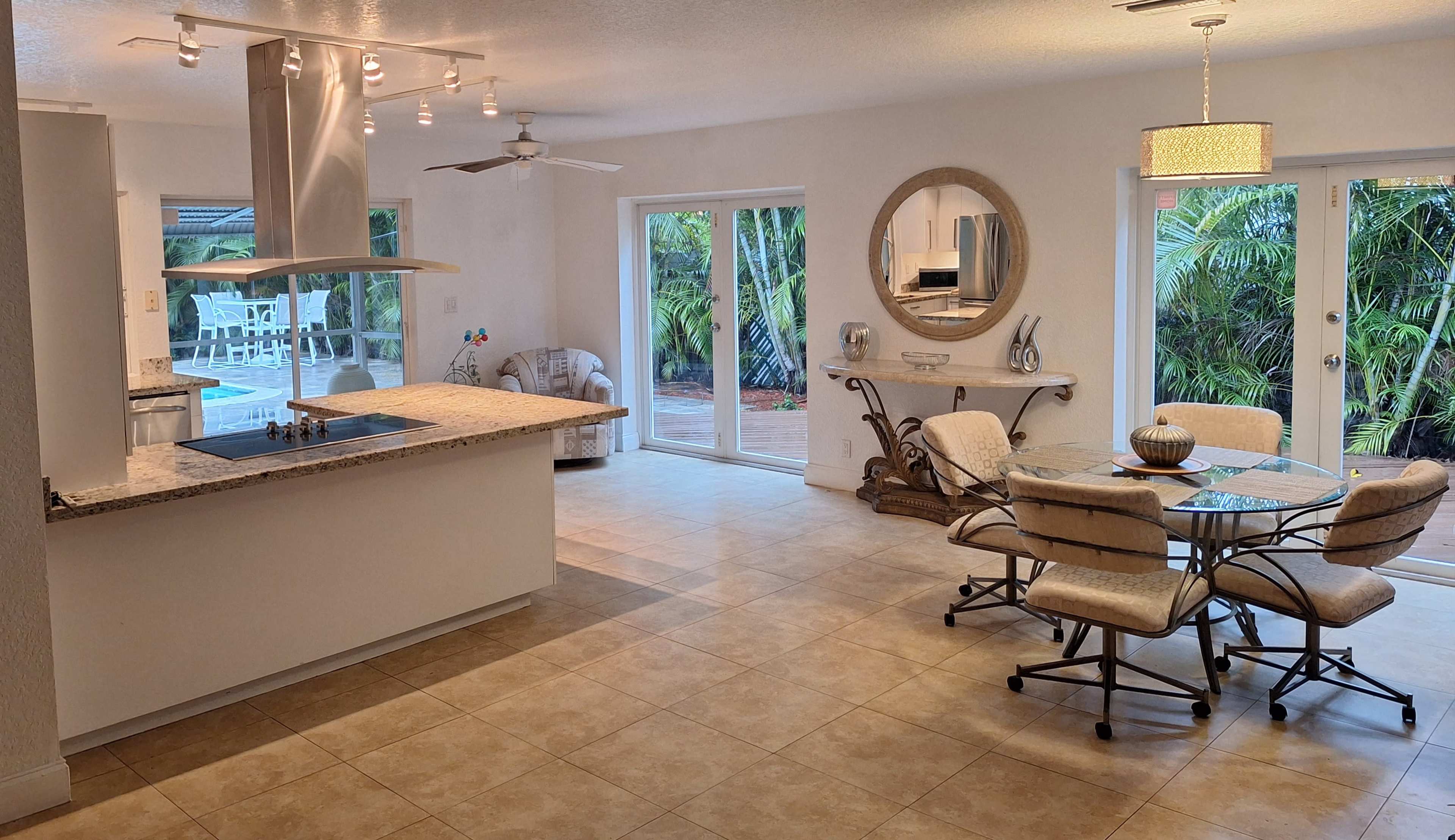 A spacious, open-concept kitchen and dining area with a central island, a round dining table, and large windows overlooking a patio surrounded by greenery.