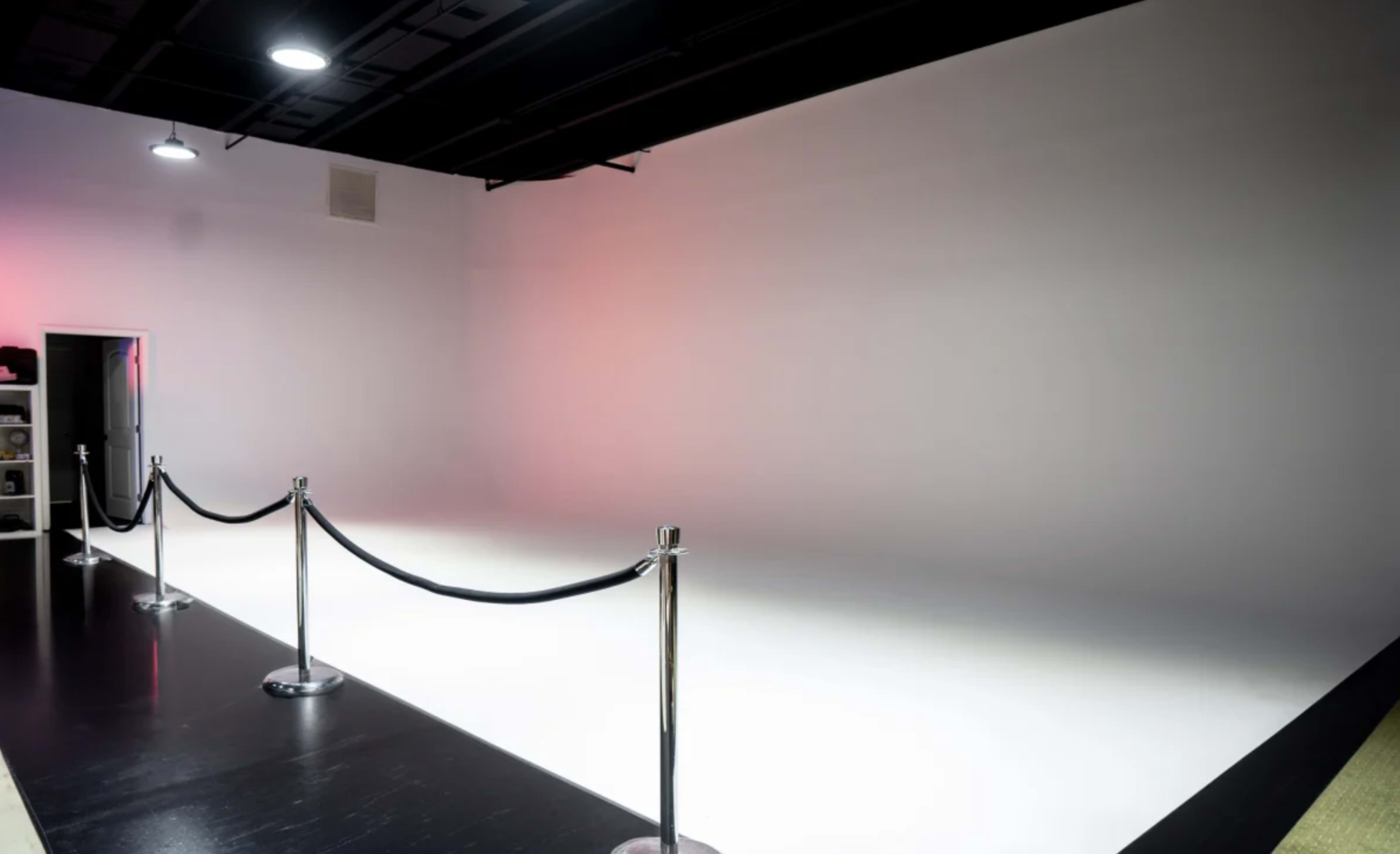 The image shows an empty studio space with a white backdrop and black flooring, separated by velvet ropes.