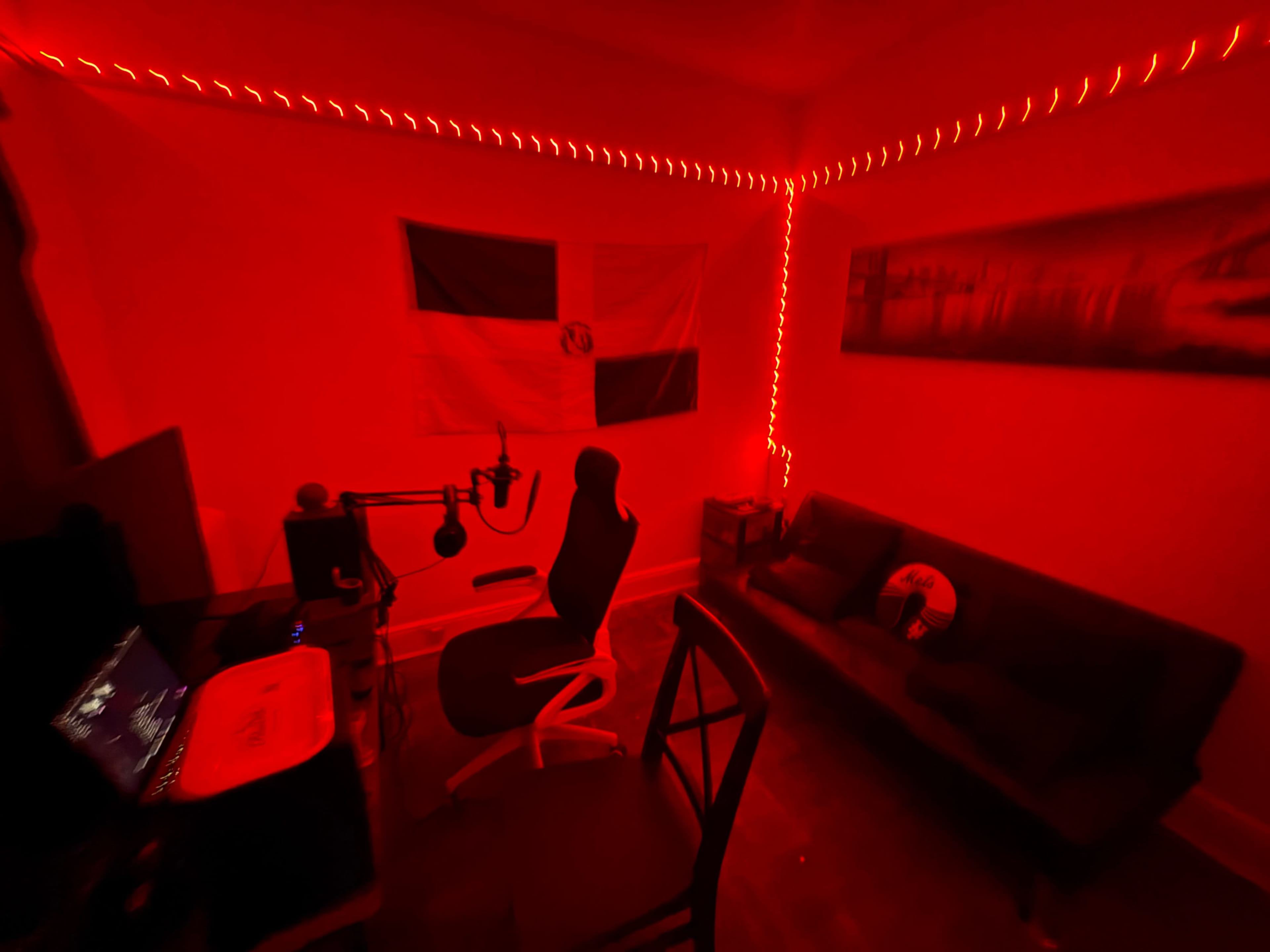 The image shows a dimly lit room with red lighting, featuring a microphone on a stand, a gaming chair, a couch, and a wall adorned with a flag.