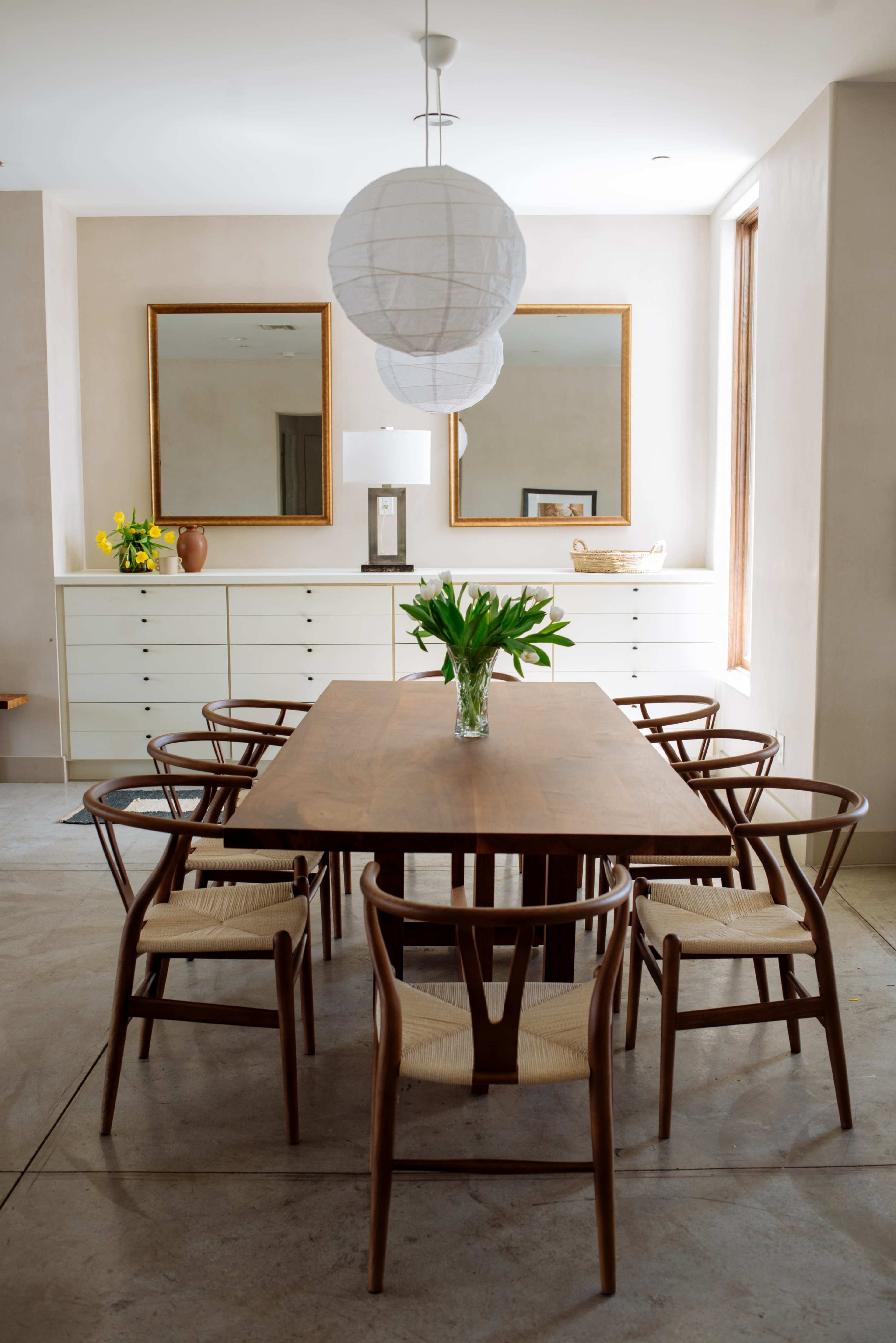A modern dining room features a wooden table surrounded by eight chairs, with two large mirrors and a pendant light above, and a vase of flowers on the table.