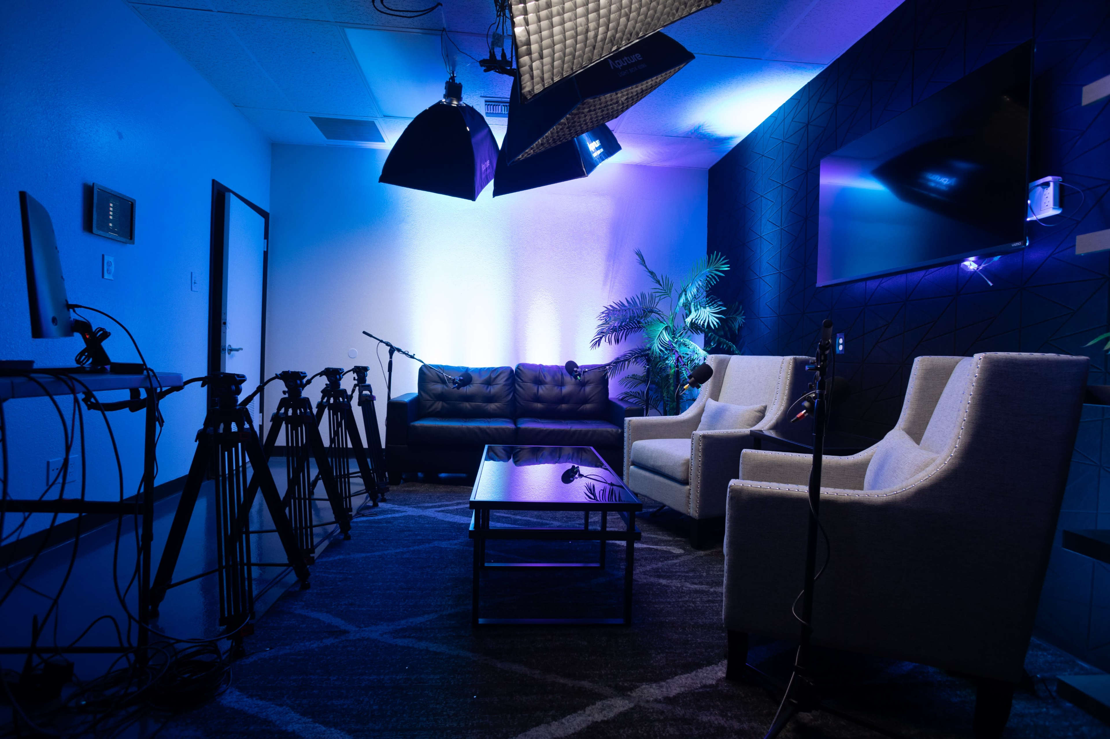 A modern studio setup featuring a black couch, two white chairs, a coffee table, and multiple cameras arranged in a well-lit space with blue and purple lighting.