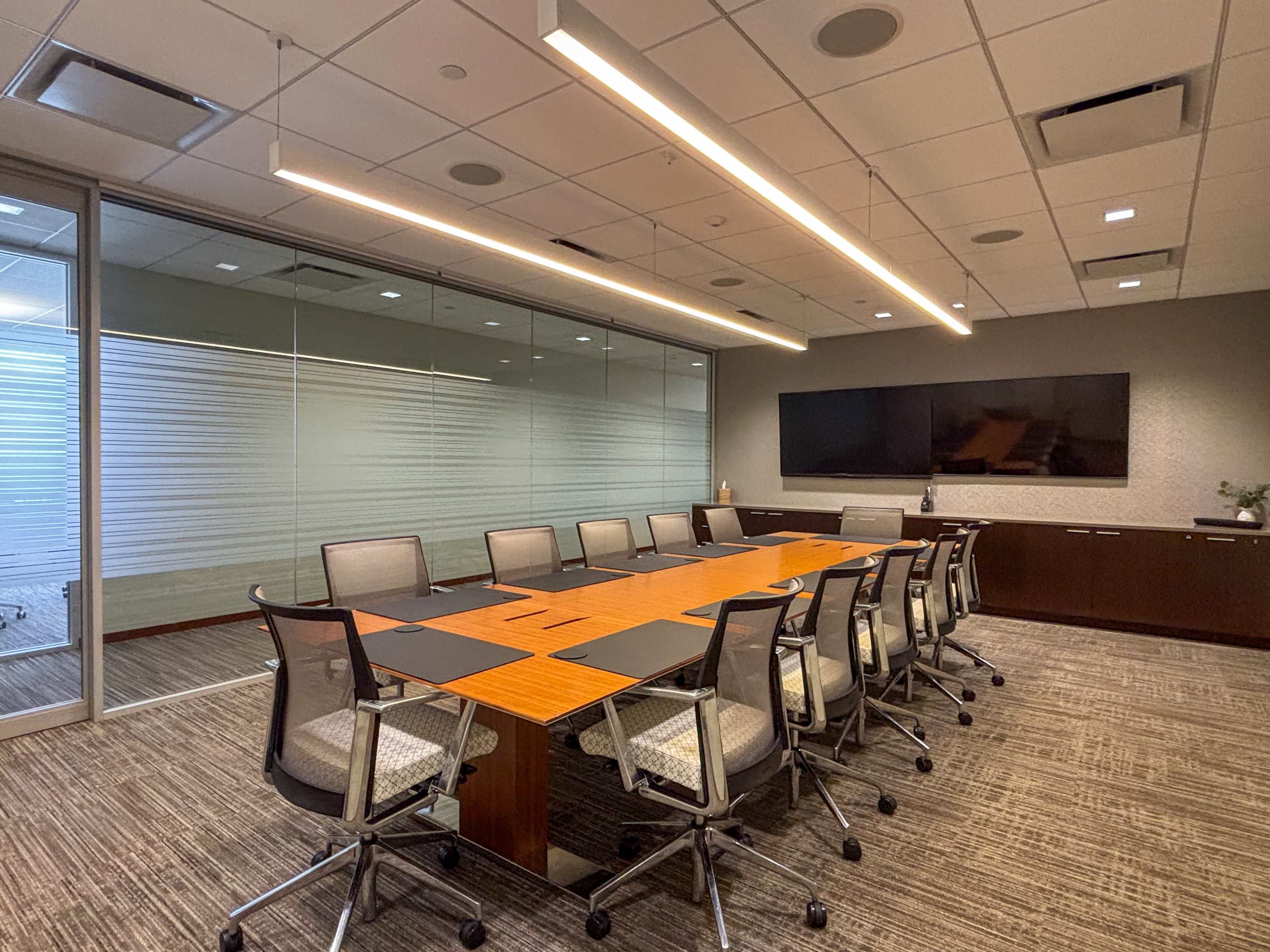 A modern conference room features a large wooden table surrounded by ergonomic chairs, with a wall-mounted screen and glass walls allowing natural light.