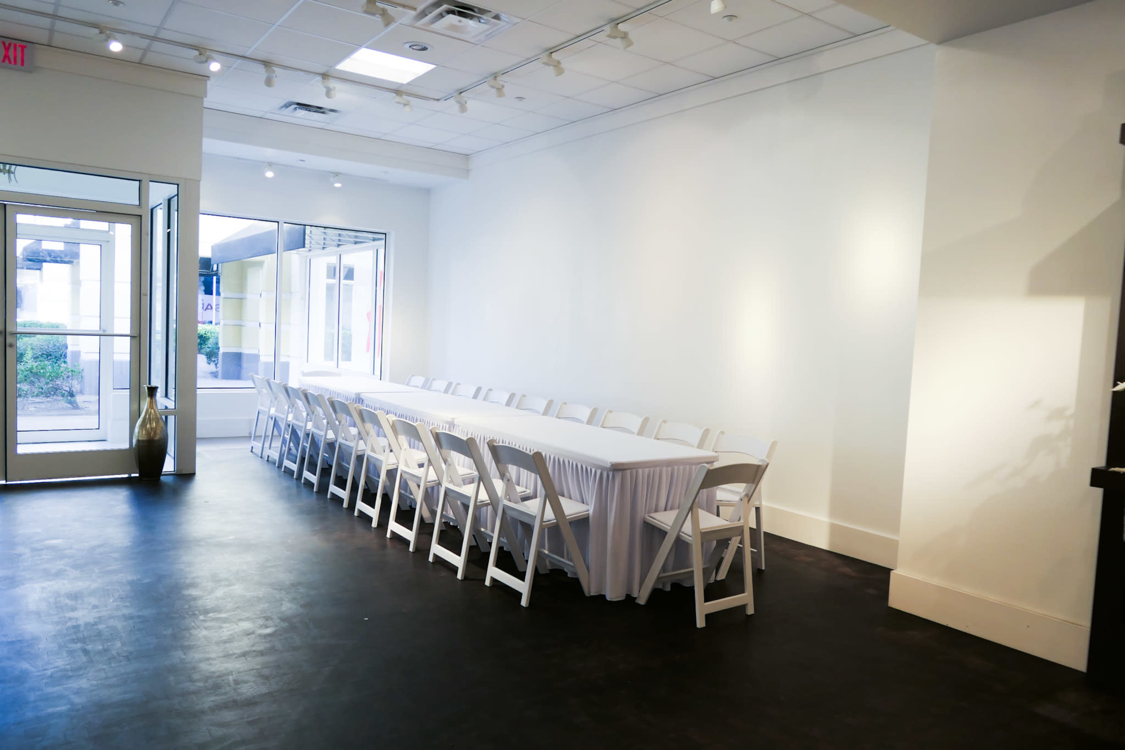 The image shows a spacious room with a long white table covered by a tablecloth, surrounded by white folding chairs, and large windows letting in natural light.