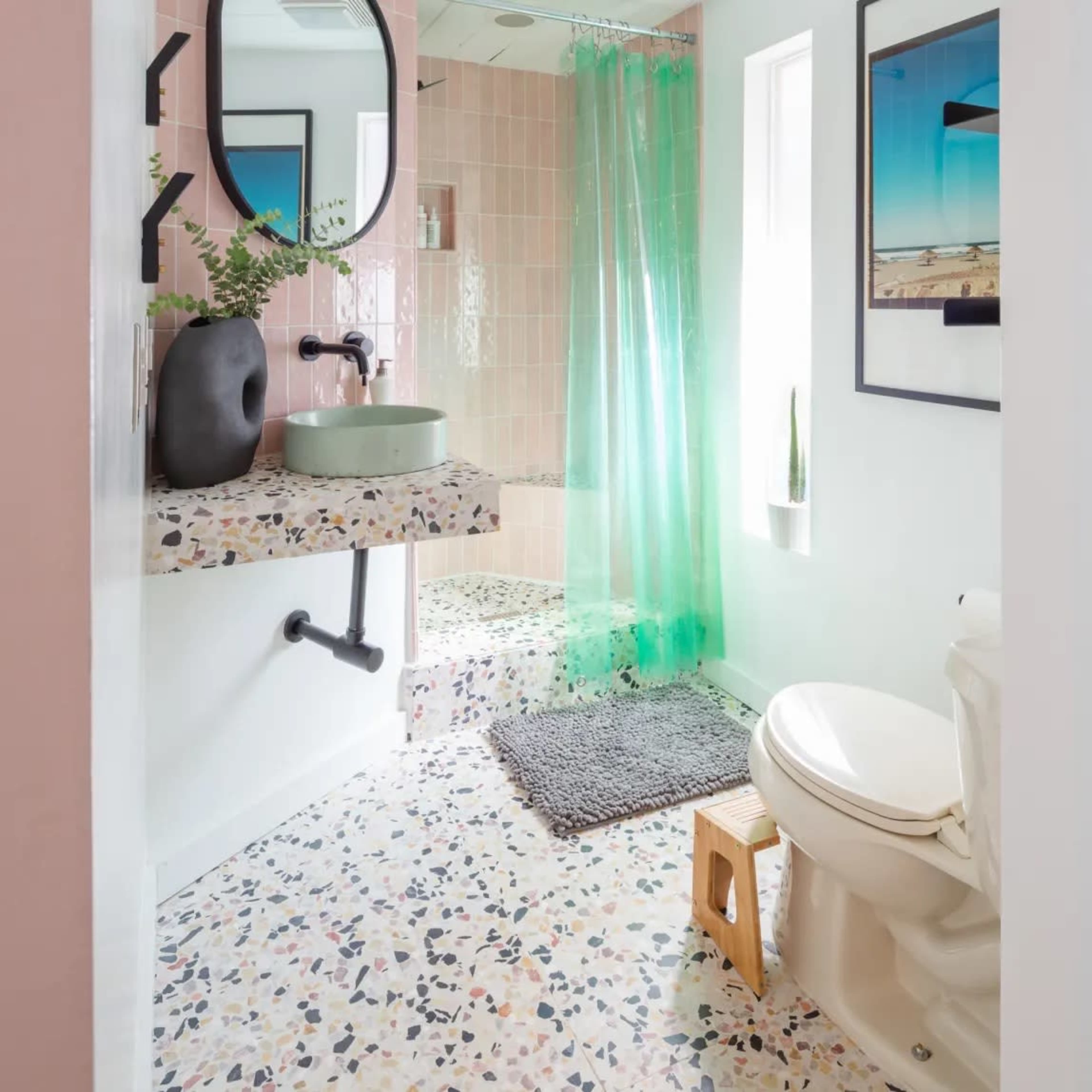 A modern bathroom featuring a circular mirror, a green shower curtain, a sink with a round basin, and a toilet beside a terrazzo floor with colorful speckles.