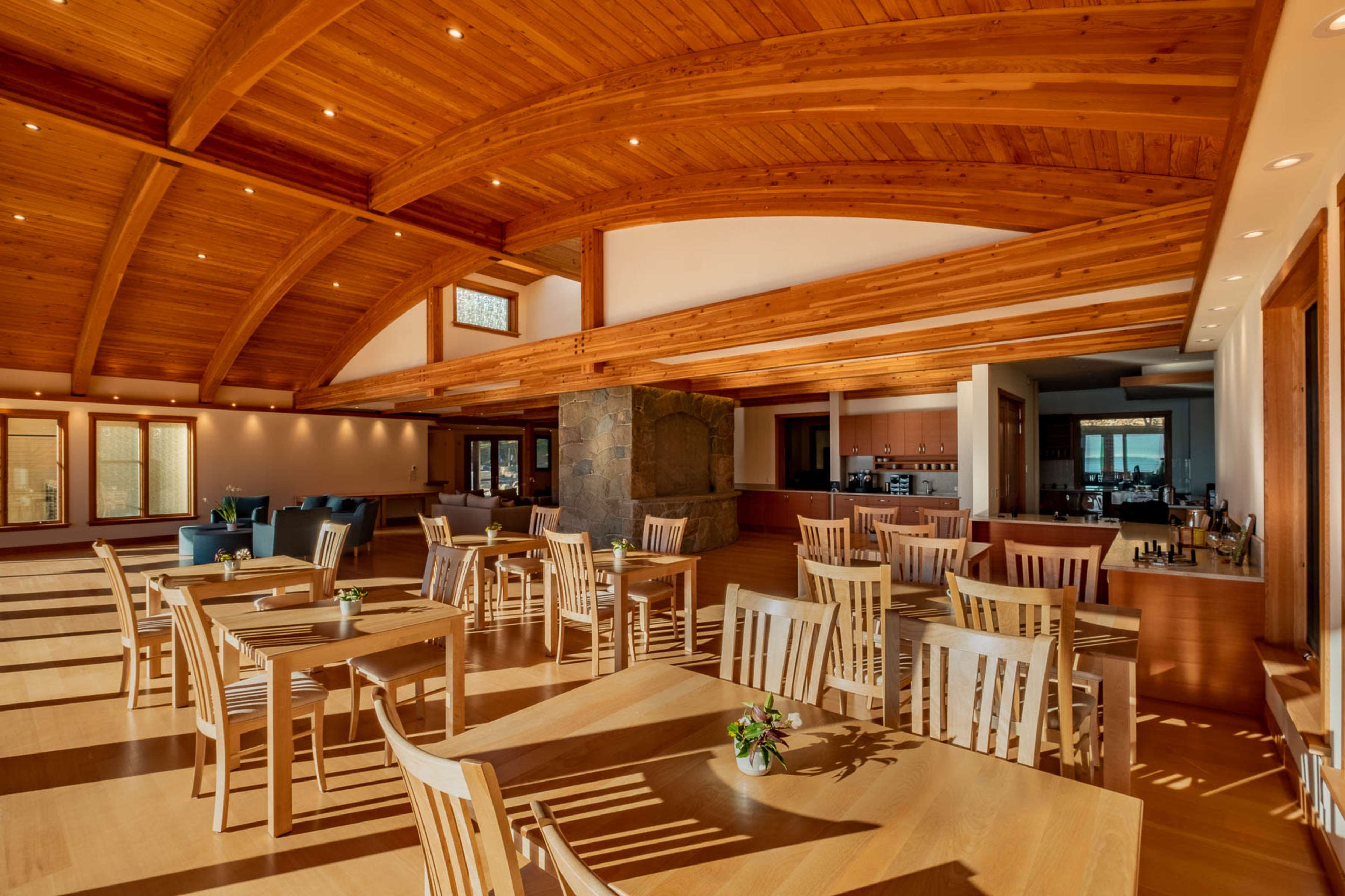A spacious dining area with wooden tables and chairs, featuring a high wooden ceiling and large windows that allow natural light to fill the space.