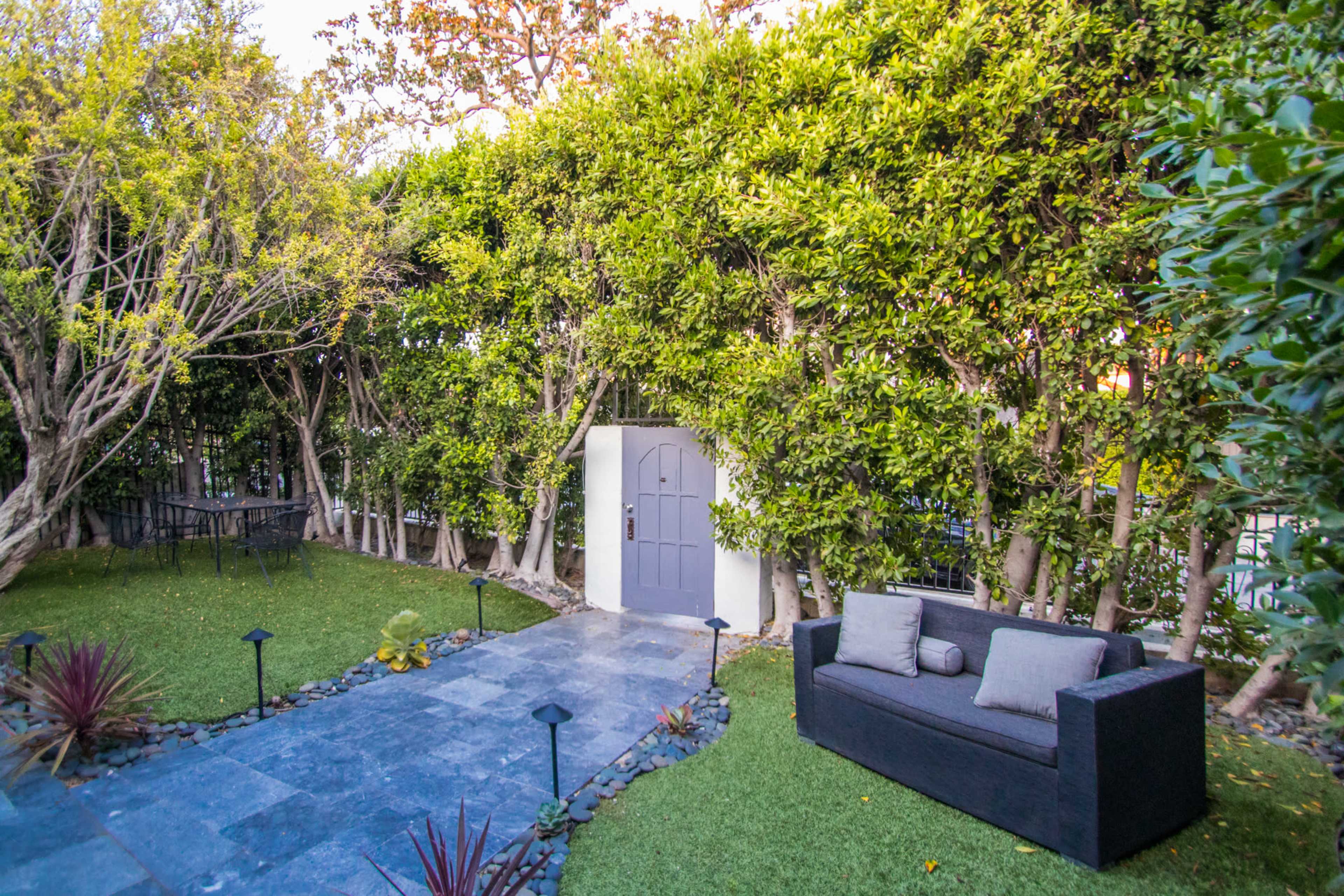 The image shows a neatly landscaped garden with a stone pathway leading to a gray door, flanked by lush greenery and a black sofa positioned on the grass.