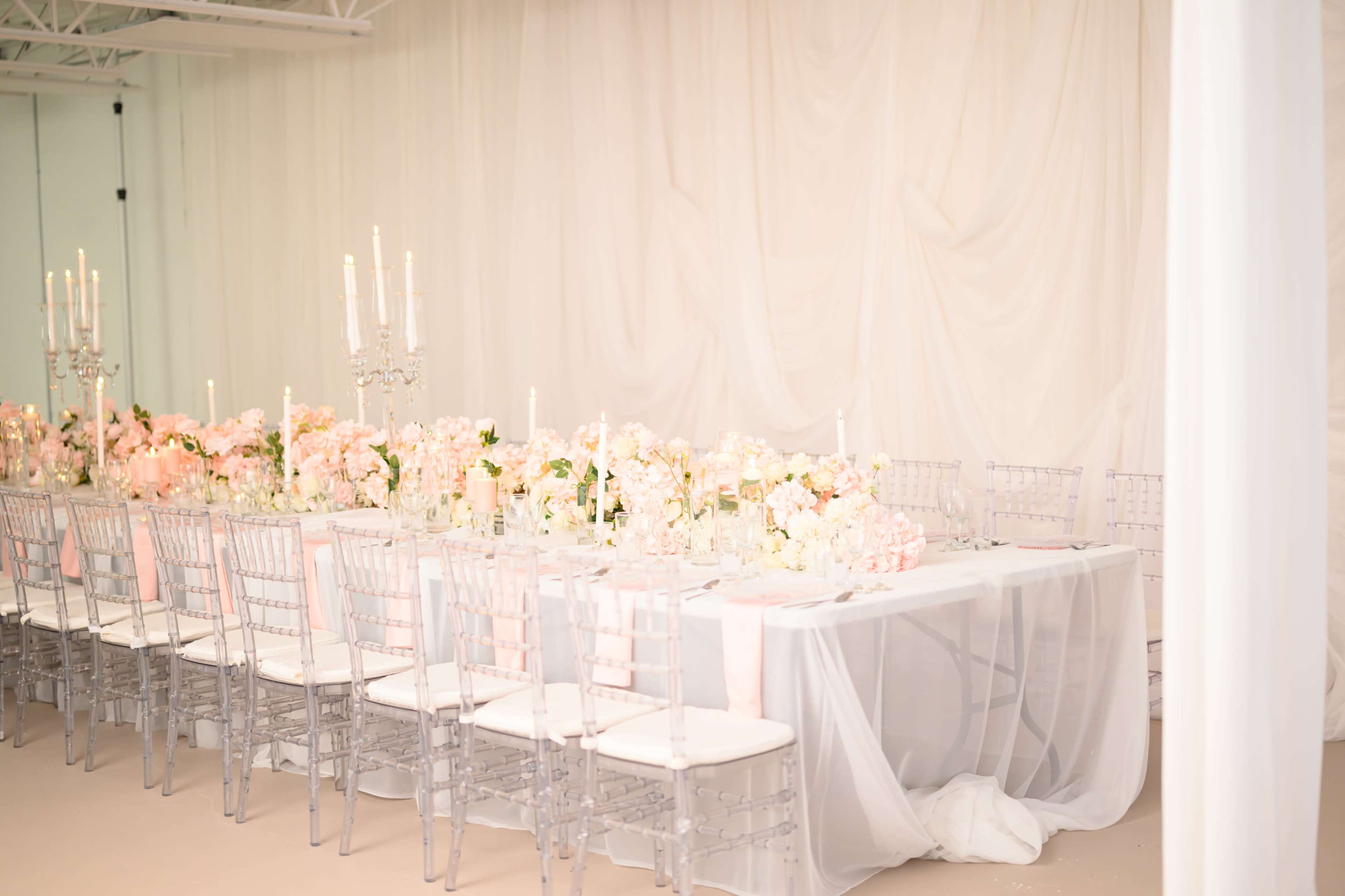 A long banquet table is set up with floral arrangements and candelabras, surrounded by clear plastic chairs in a softly draped room.