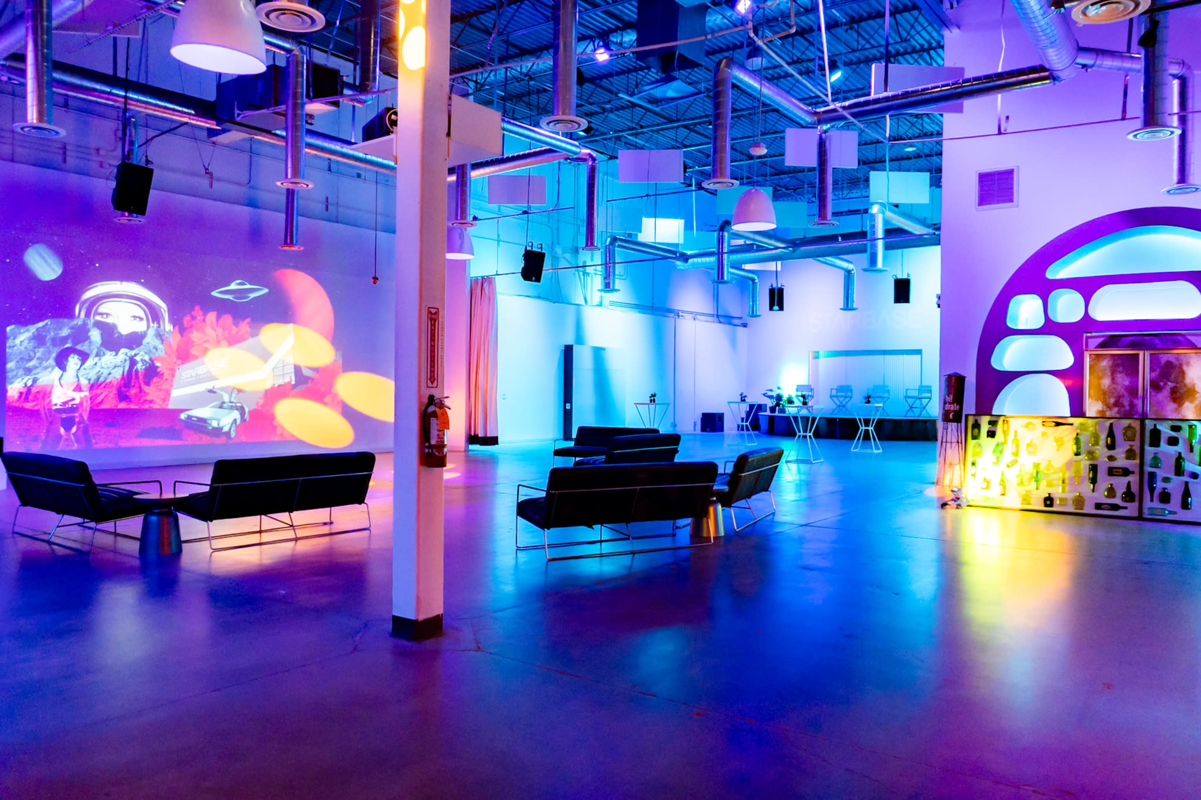 The image shows a spacious, modern event venue with colorful lighting, featuring seating areas and a large projection screen displaying vibrant visuals.