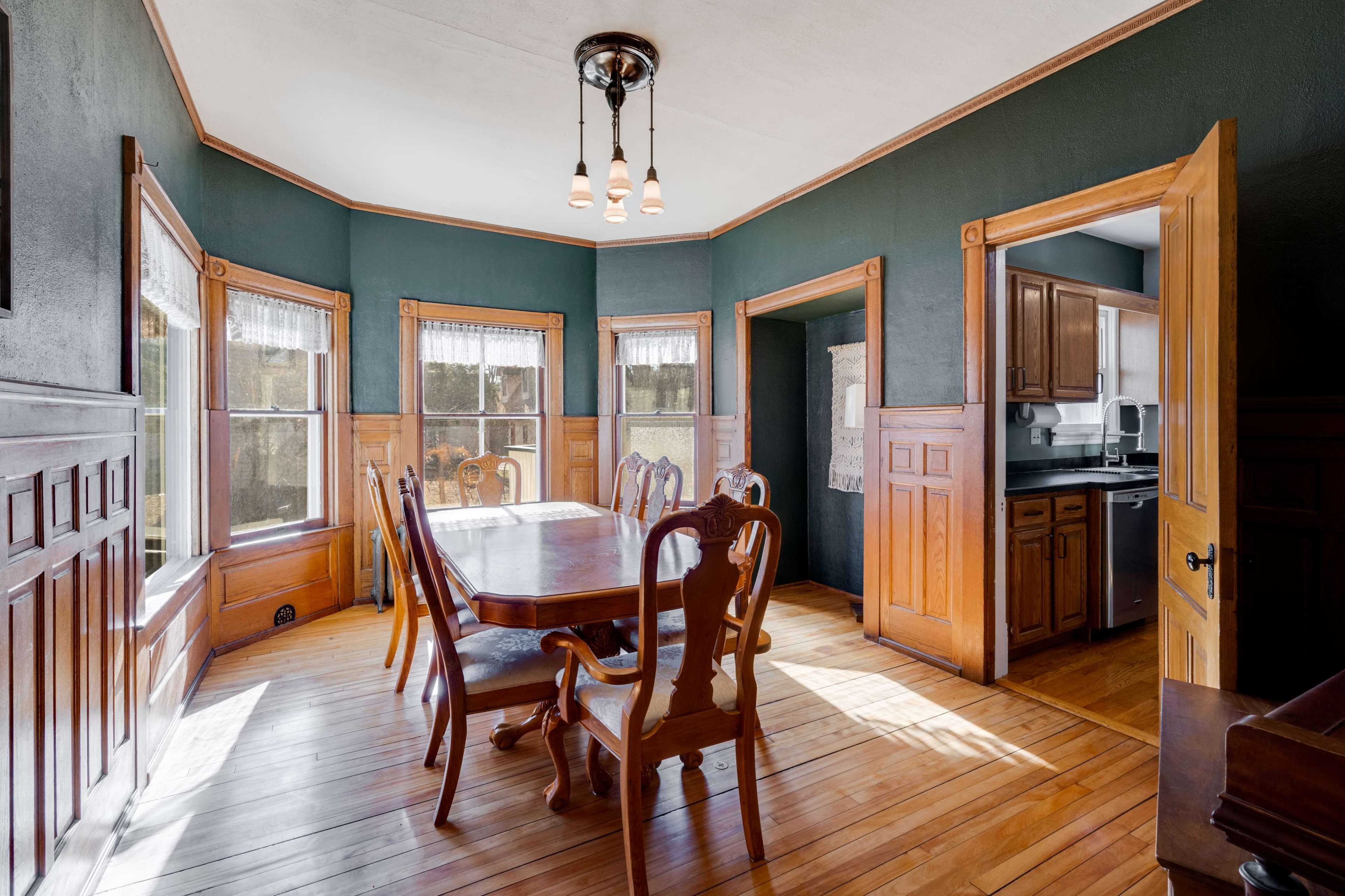 A sunlit dining room with a large table surrounded by wooden chairs, featuring dark green walls and wooden trim.