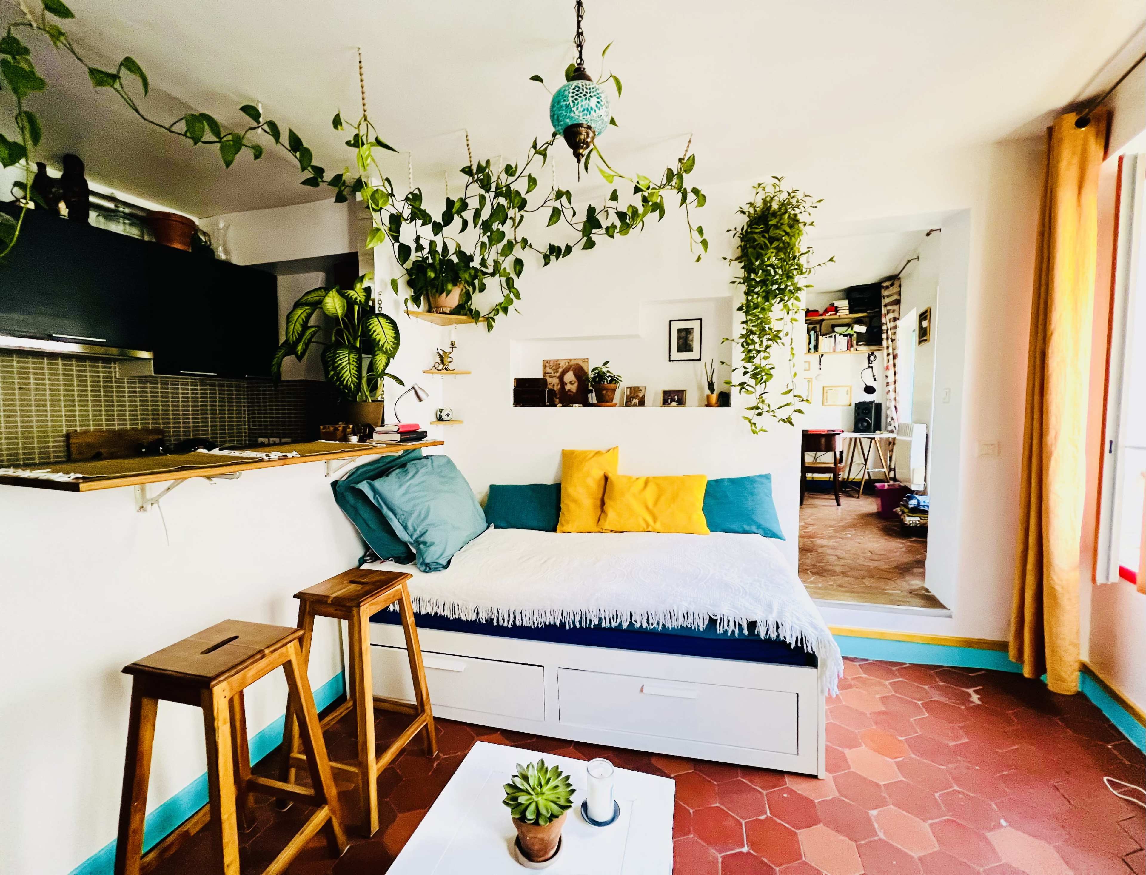 The image depicts a cozy living space featuring a daybed with colorful cushions, potted plants hanging from the ceiling, and wooden stools beside a light-colored coffee table.