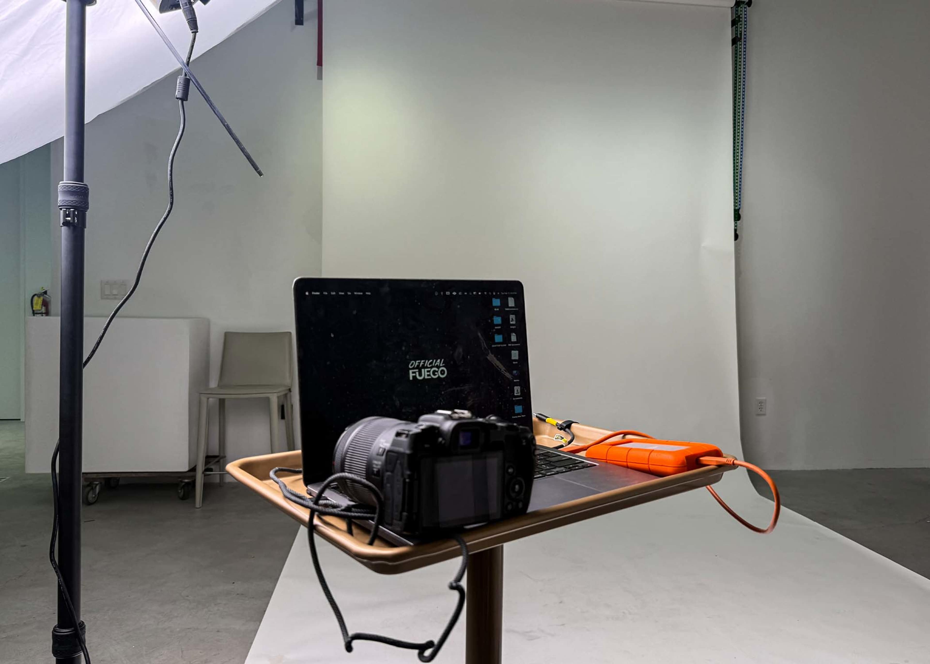 A camera and a laptop are set on a table in a photo studio with a white backdrop and lighting equipment.