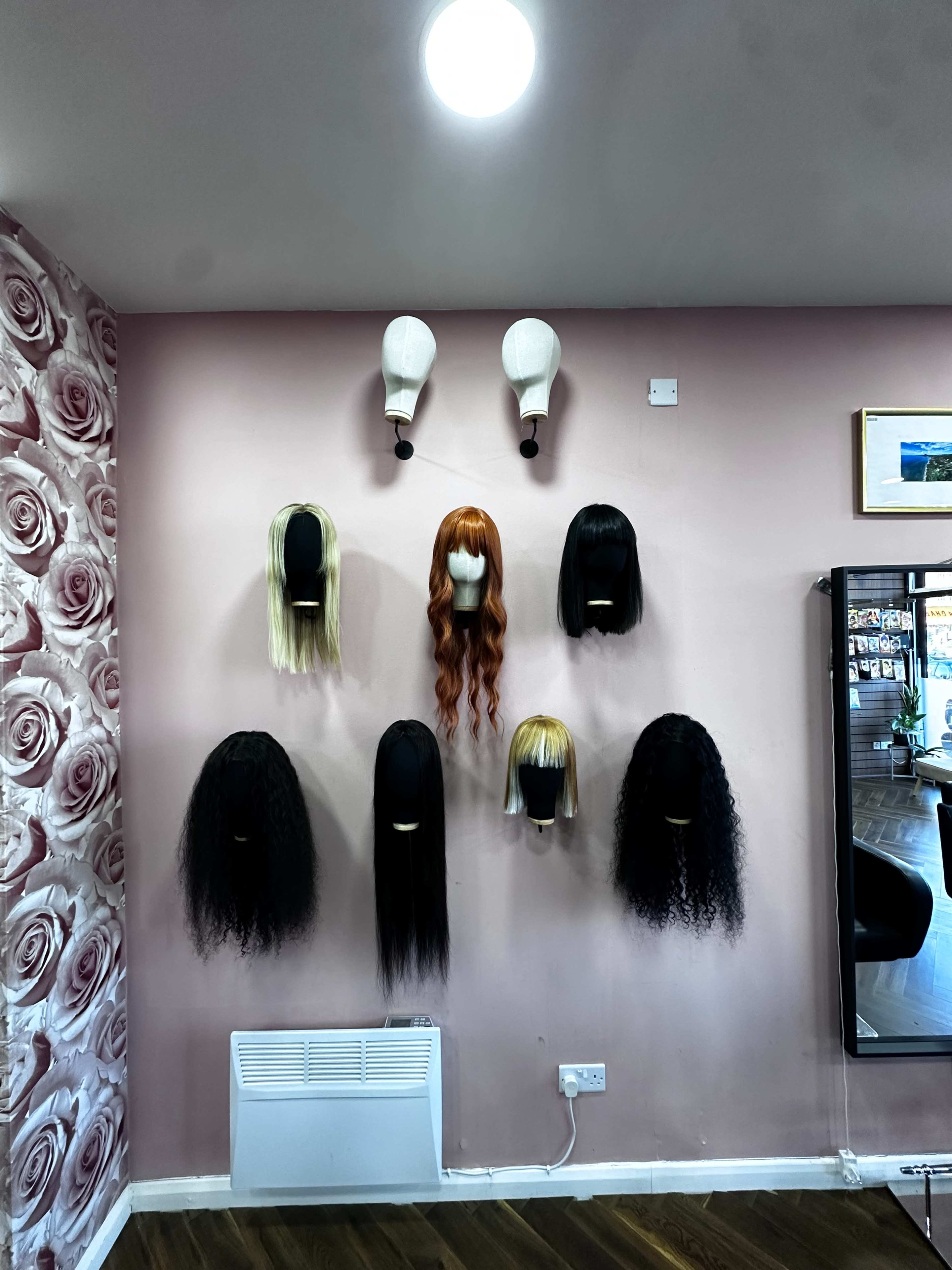 A wall displays various wigs of different styles and colors mounted on white wig stands.