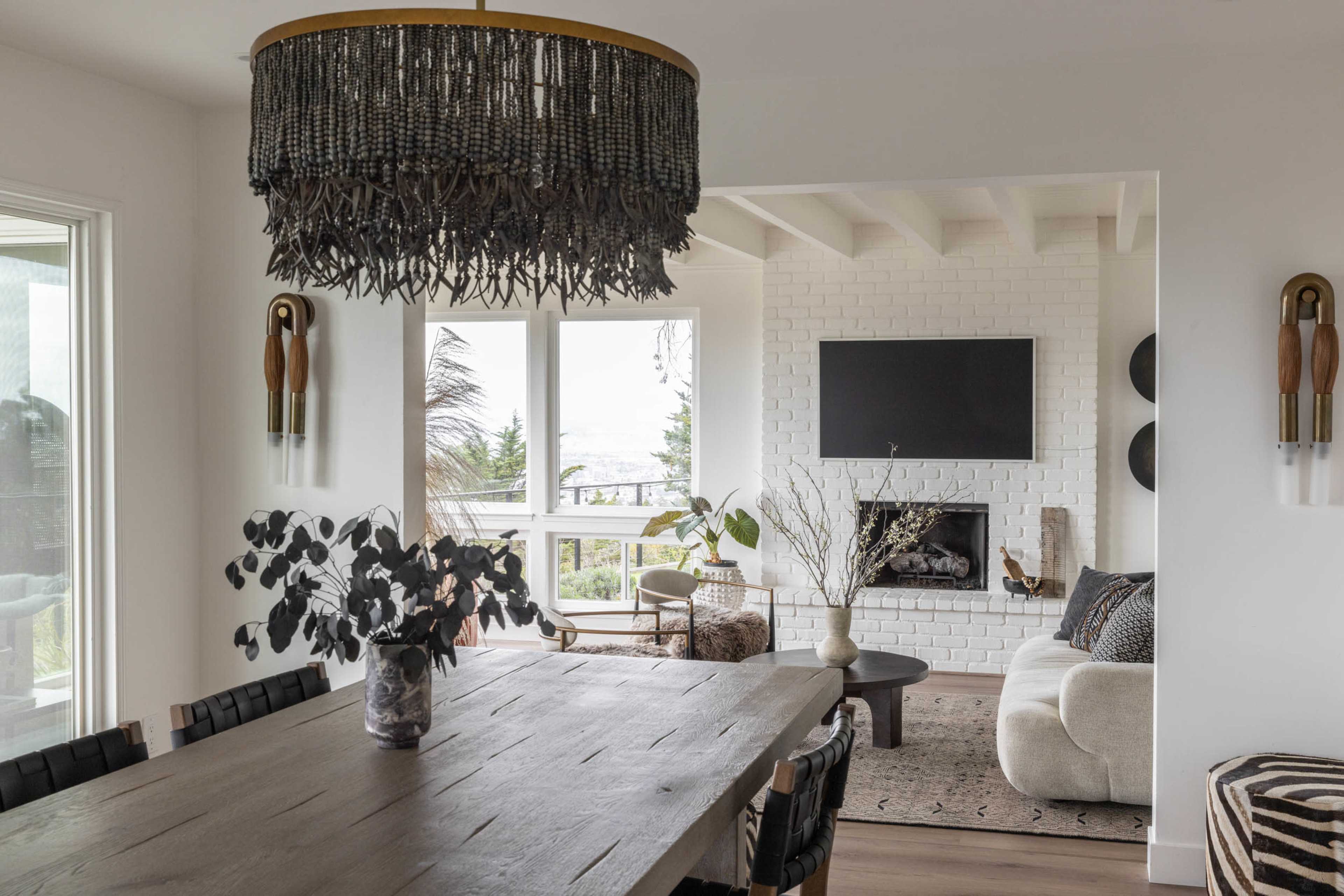 A modern dining room features a wooden table and a large chandelier, with an open view leading to a living area accented by a white brick fireplace and a mounted television.