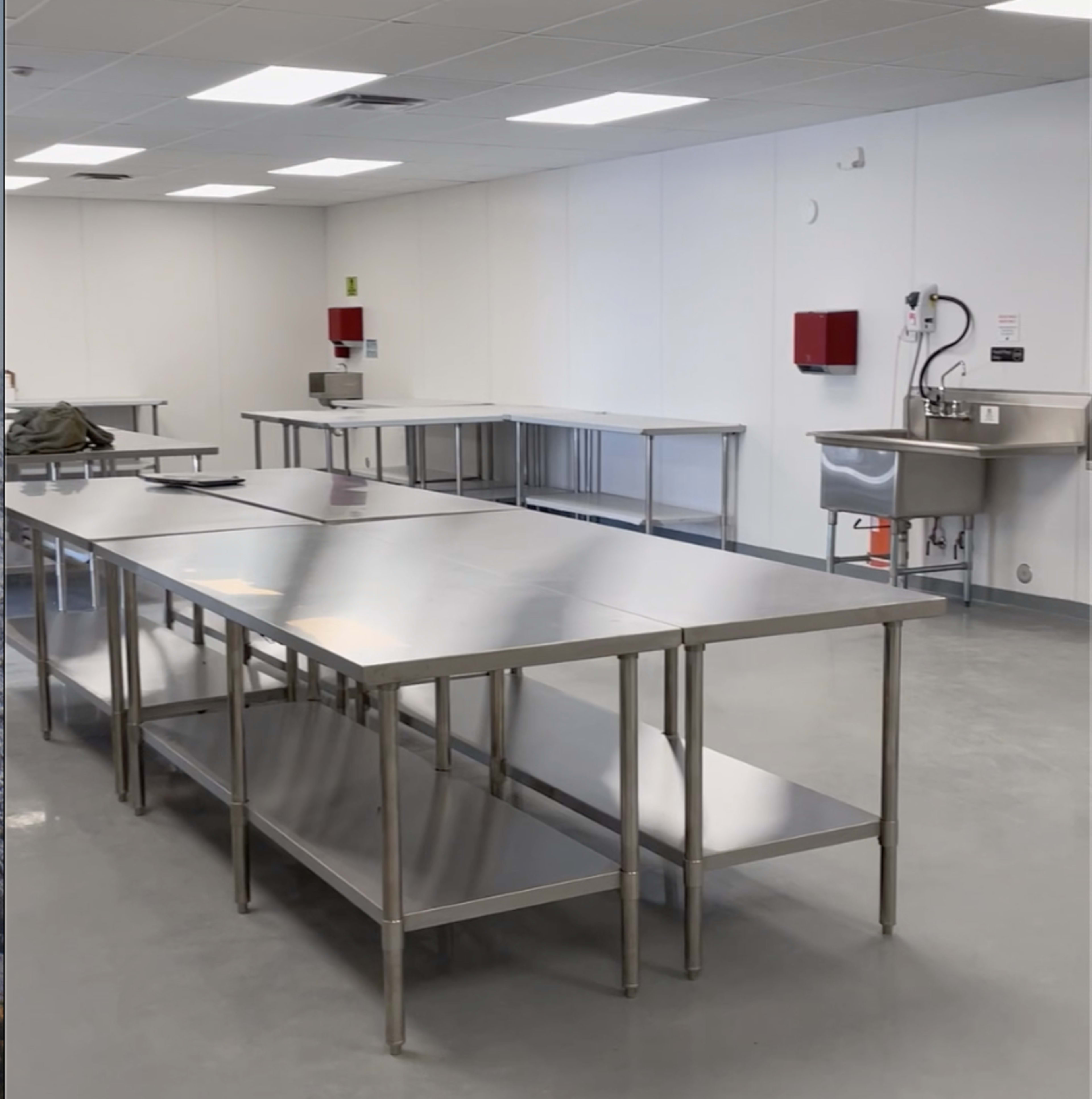 A spacious industrial kitchen with several stainless steel work tables and a sink against a white wall.