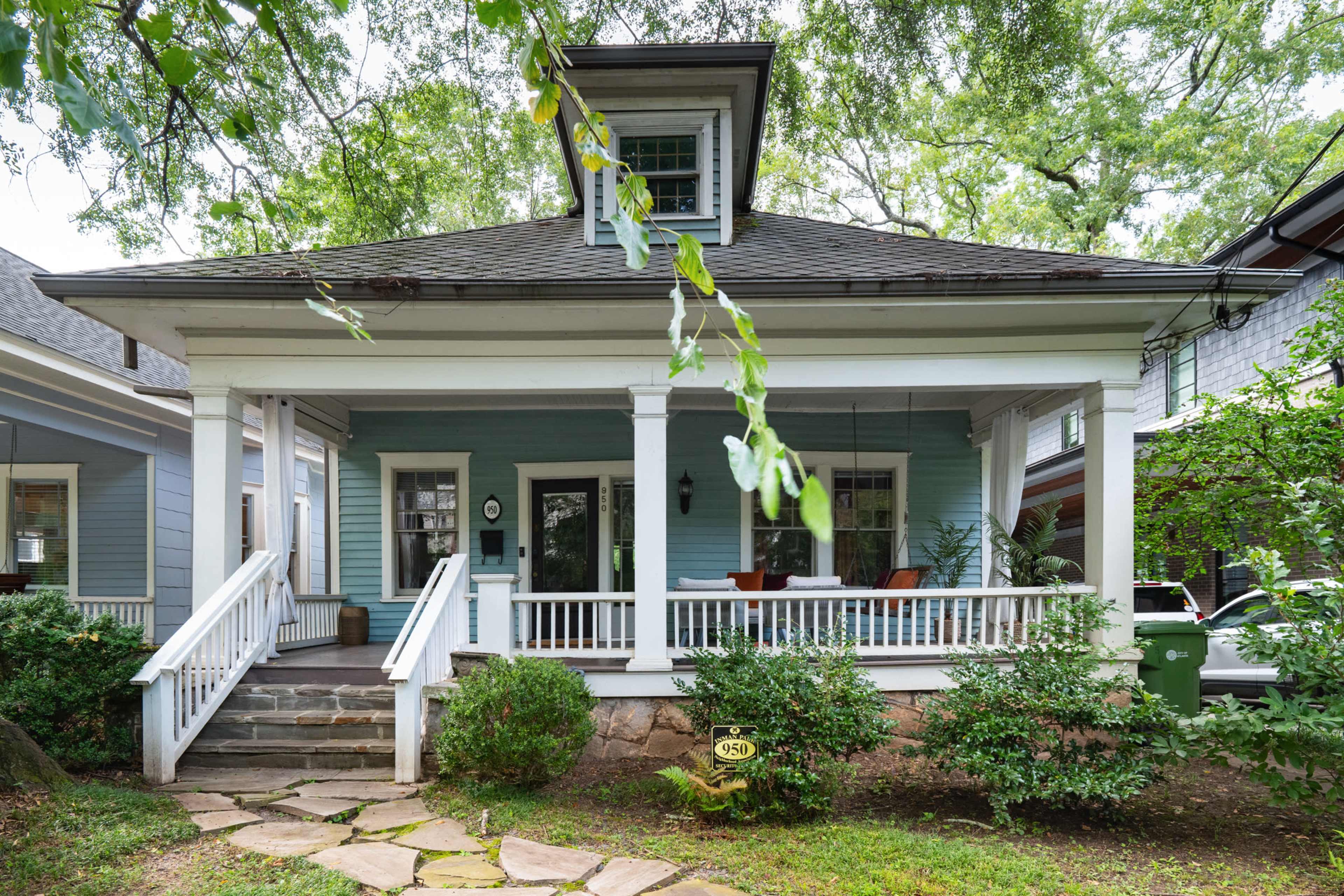 The image shows a light blue house with a covered porch, white columns, and a well-maintained garden.