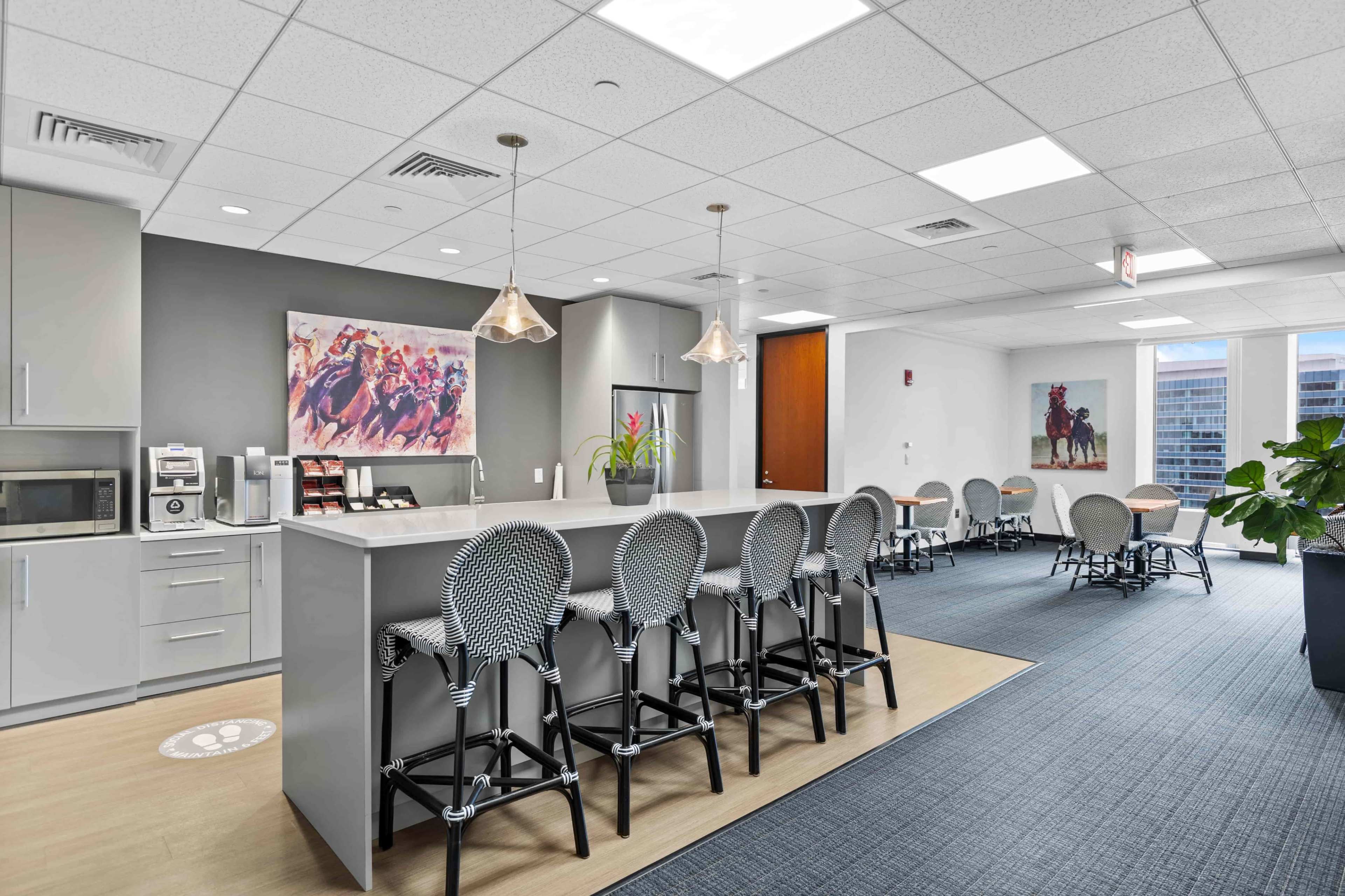 A modern office break room features a long countertop with bar stools, a coffee machine, and art on the walls.