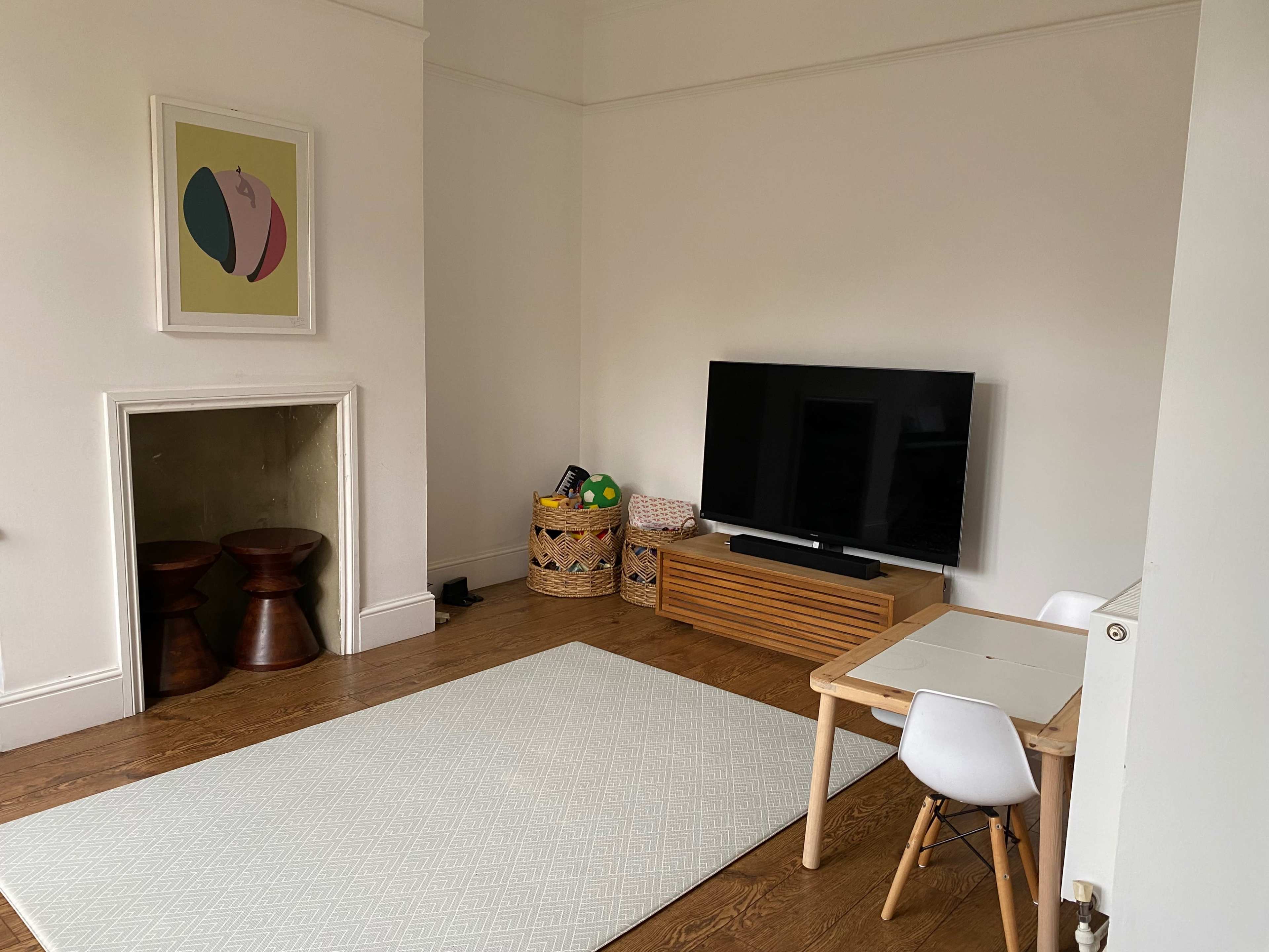 A minimalist living room features a television stand, a small wooden table with a chair, and a patterned area rug on a wooden floor.