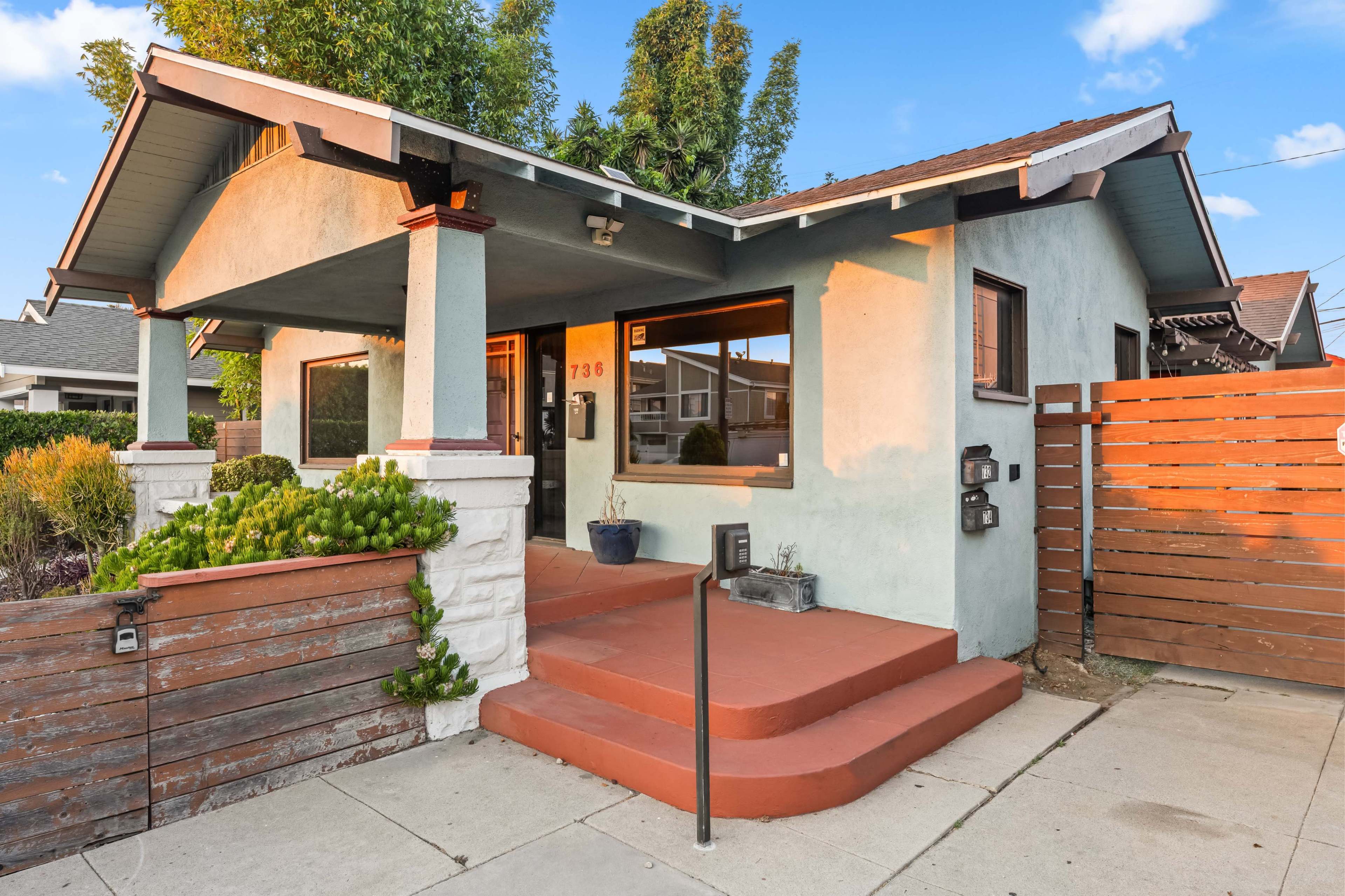 The image shows a single-story house with a covered porch, painted in light blue, and surrounded by landscaping and a wooden fence.