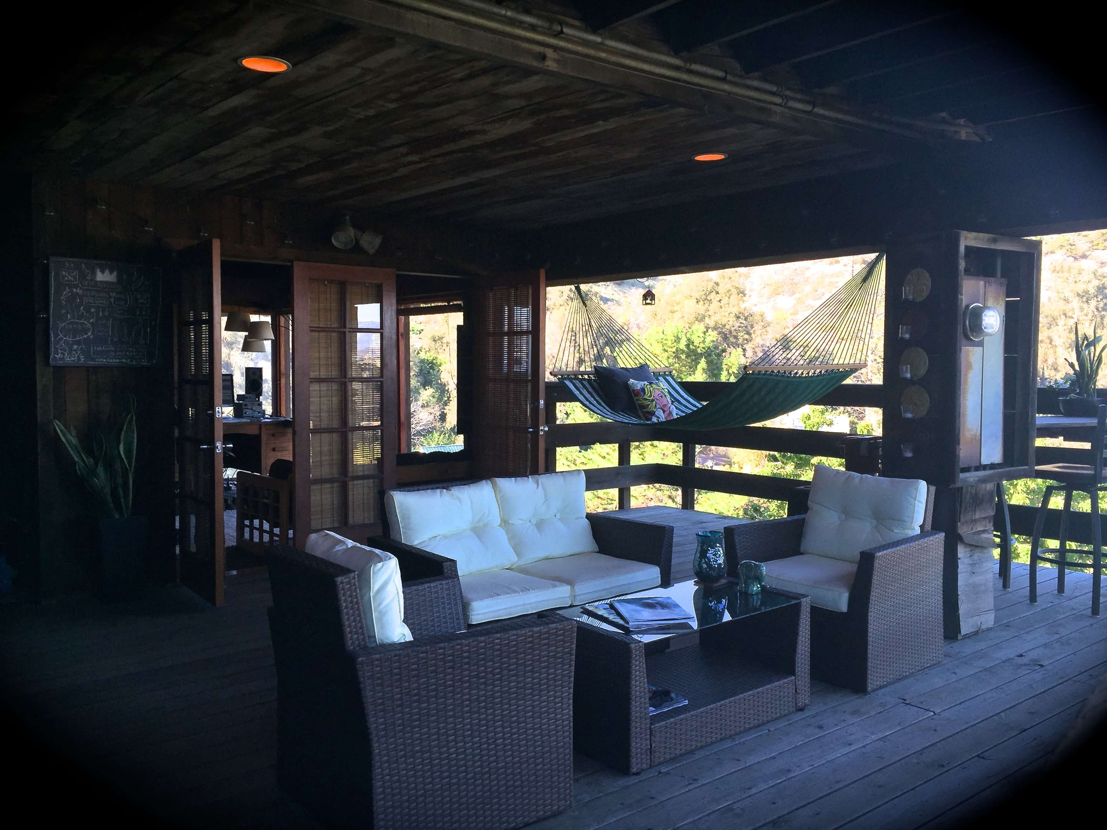 The image shows a wooden deck with seating arrangements and a hammock in a cozy outdoor setting surrounded by greenery.