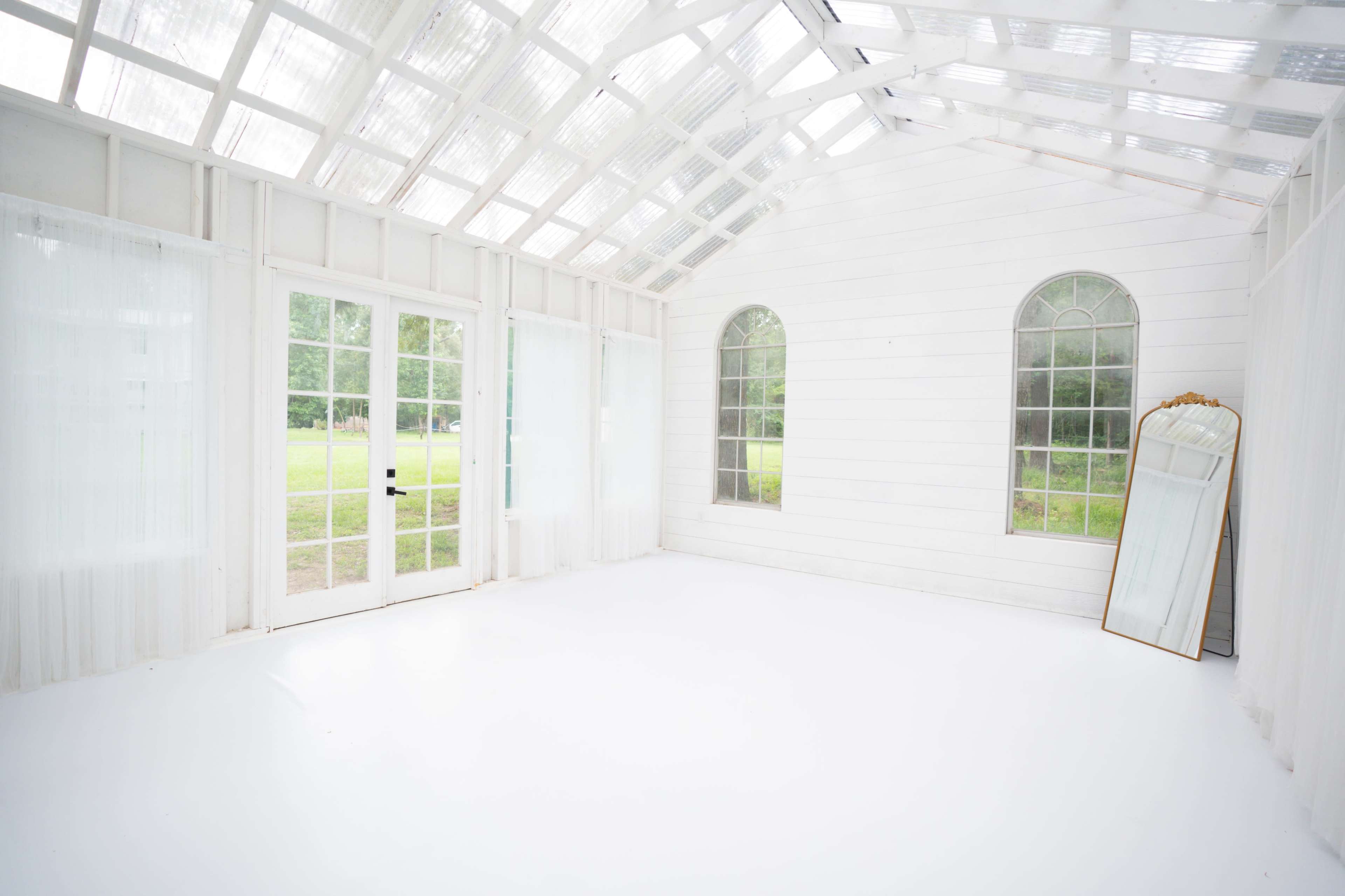 The image shows a bright, white-walled room with large windows, a glass ceiling, and a mirror leaning against the wall.
