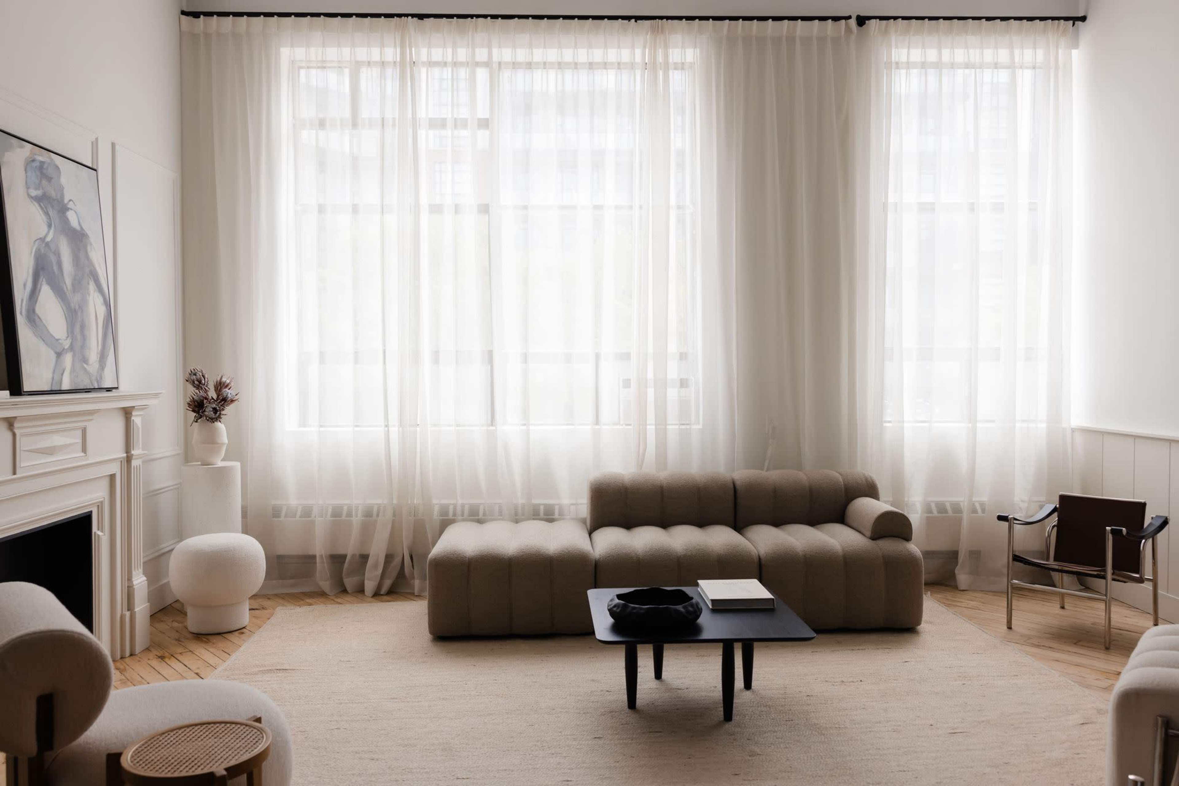 The living room features a light-colored sectional sofa, a black coffee table, and large windows dressed with sheer curtains, creating a spacious and airy atmosphere.