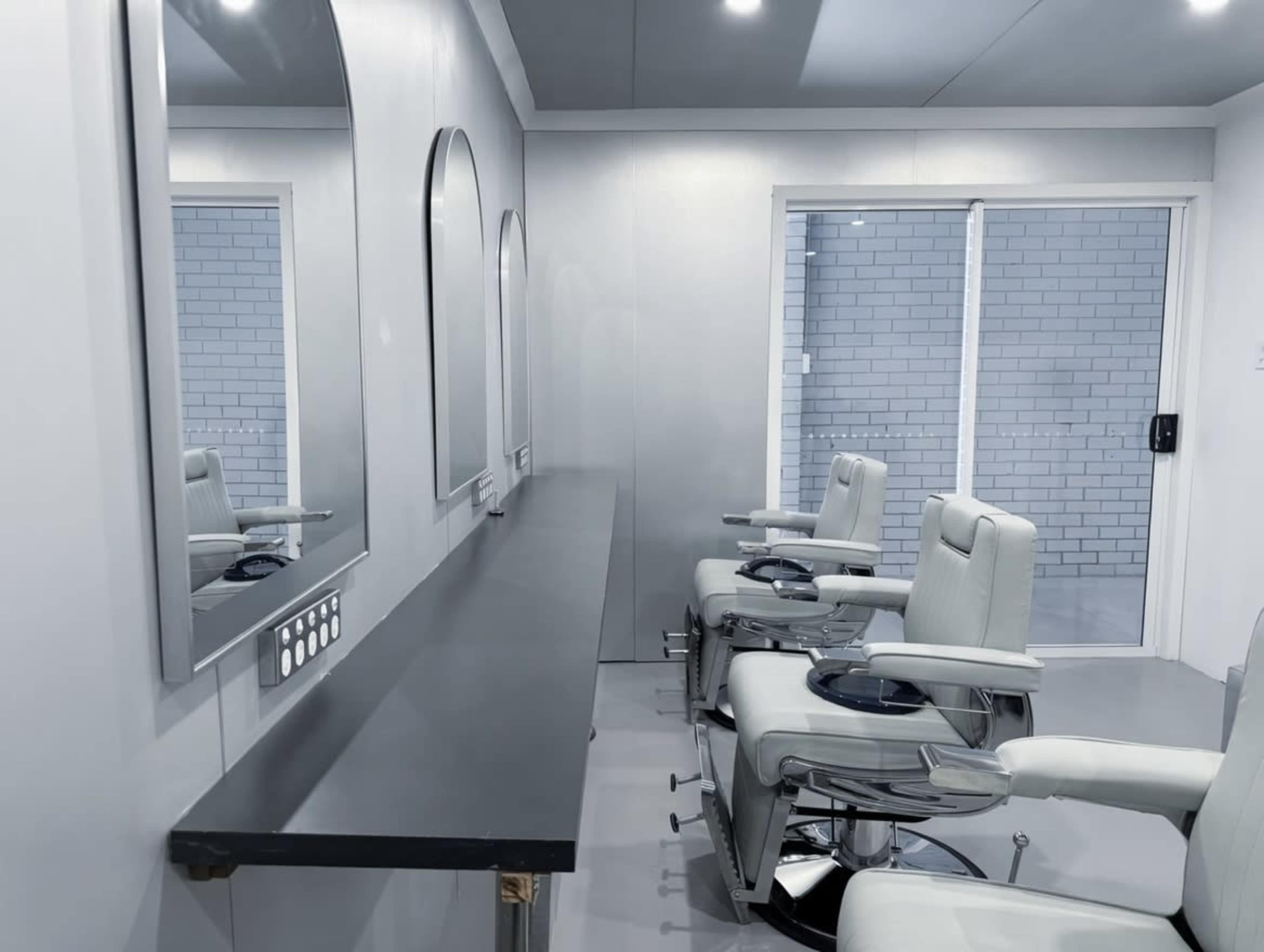The image shows a minimalist beauty salon interior with gray walls, two mirrors, and several styling chairs arranged in a line.