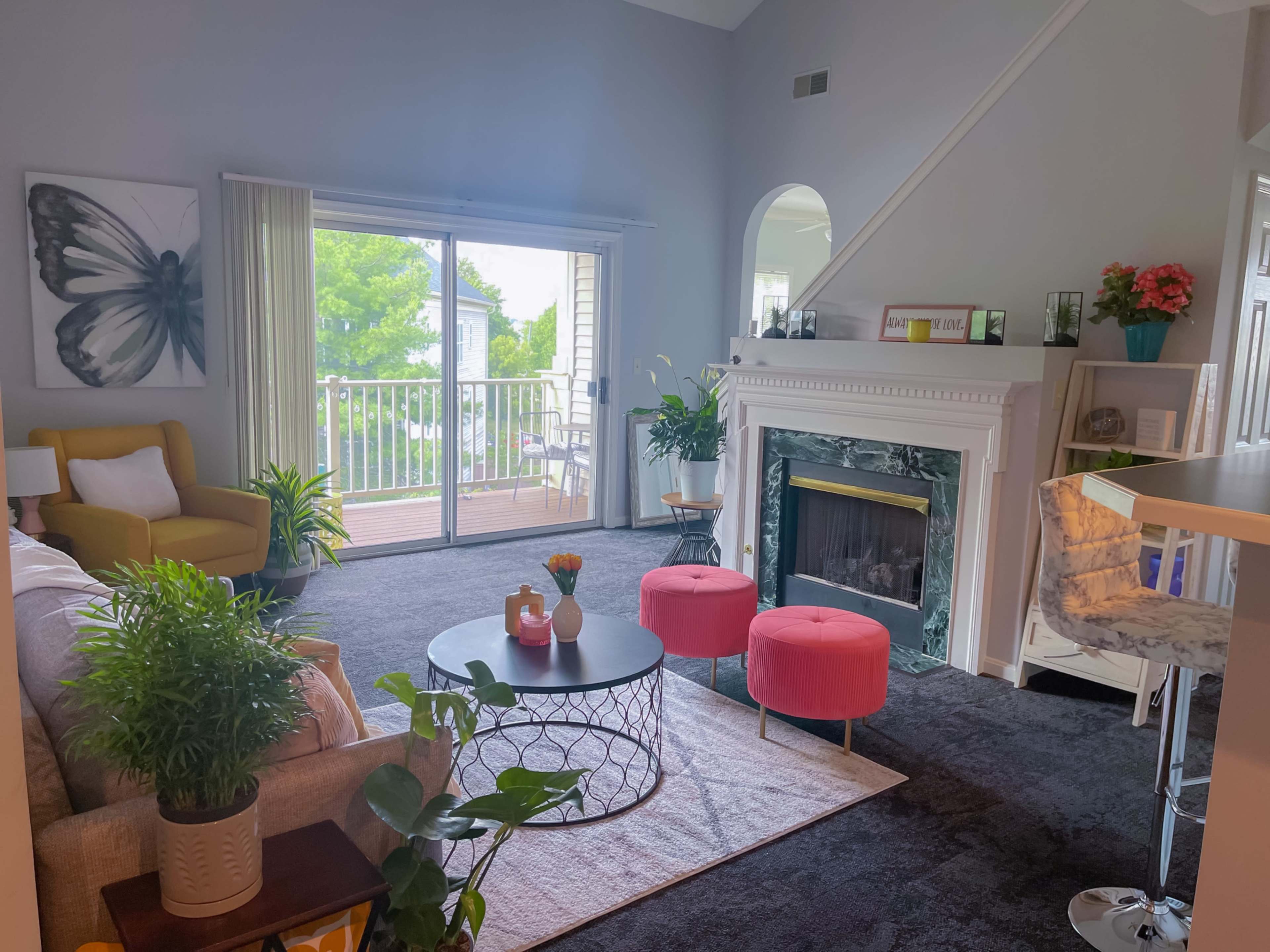 The living room features a butterfly artwork on the wall, a yellow armchair, a beige sofa, a round coffee table, and pink ottomans, with large windows offering a view of a balcony and greenery outside.