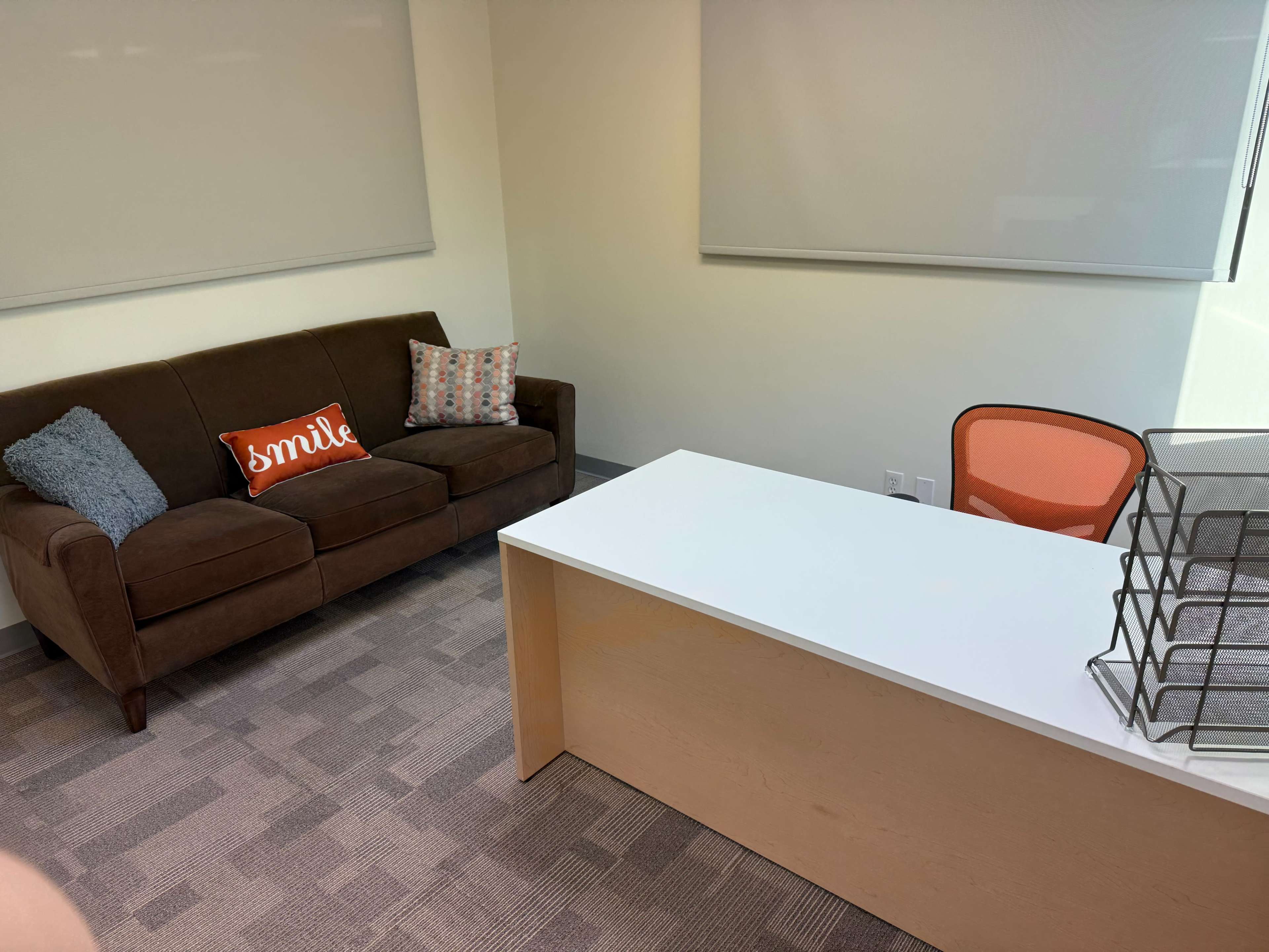 The image shows a modern office space featuring a brown couch with decorative pillows, an orange chair, and a white desk with a wire organizer.