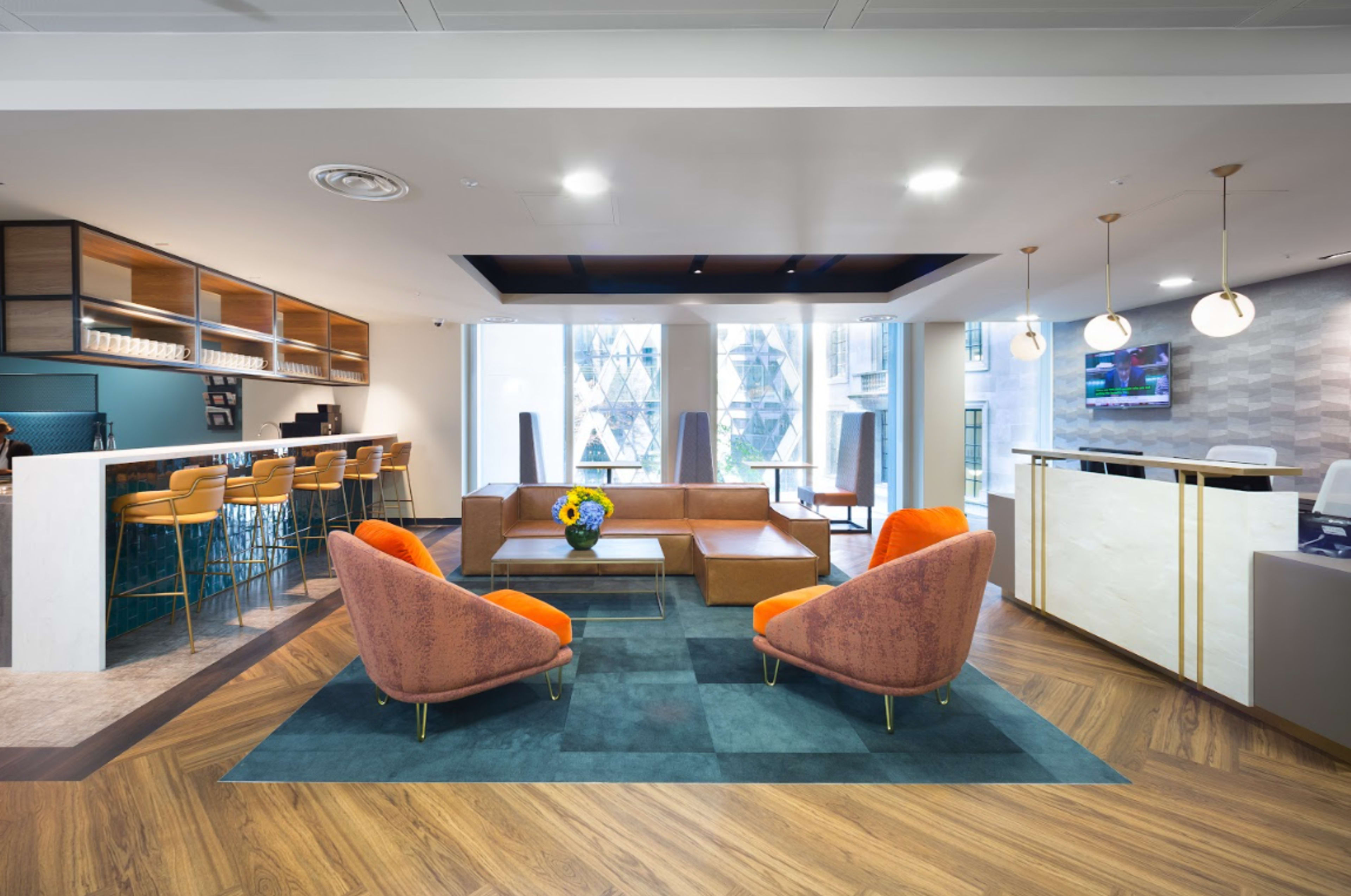 The image shows a modern office lobby with a seating area featuring orange and brown chairs, a small coffee table, and a reception desk in a bright space with large windows.