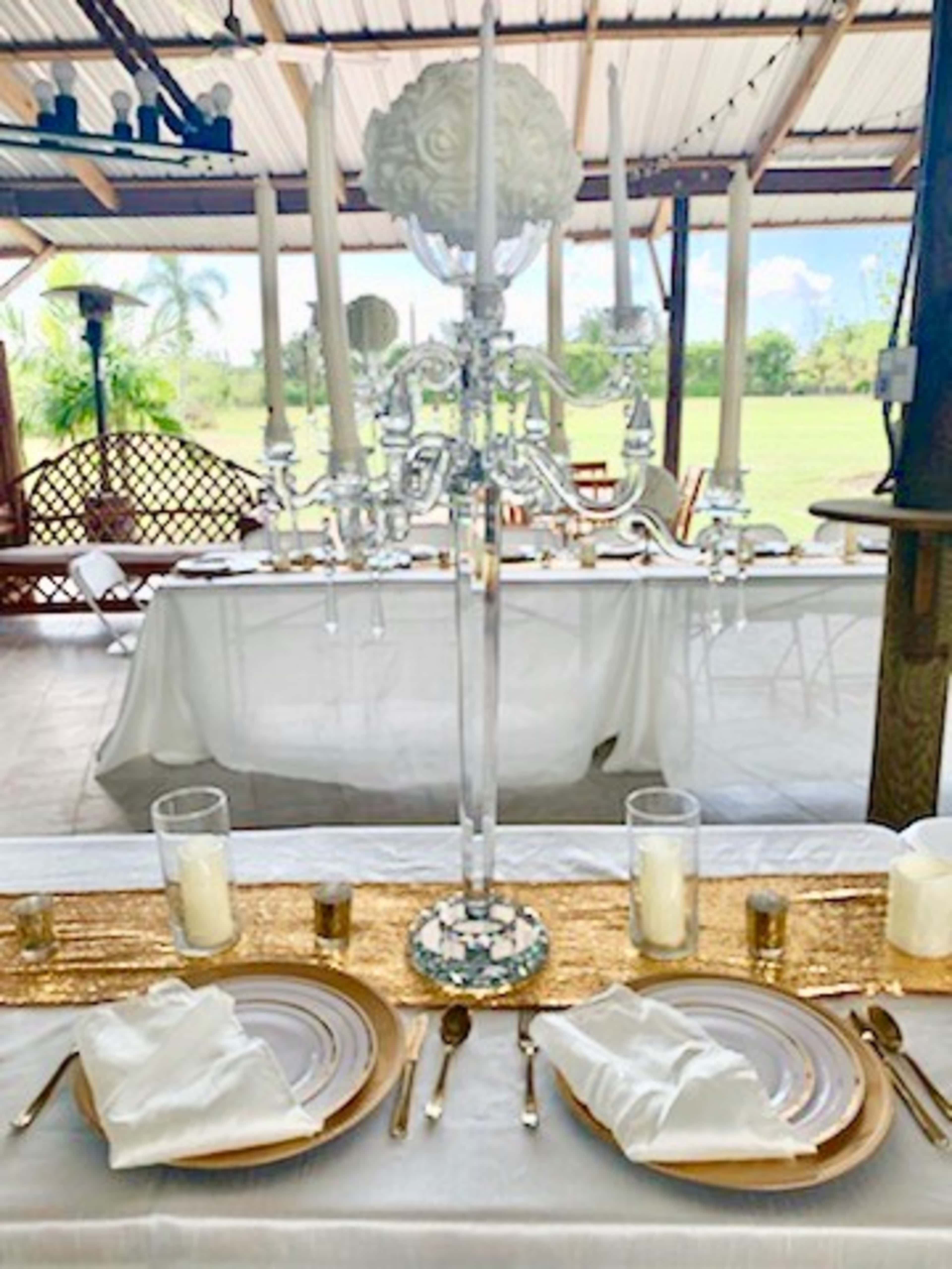 A formal dining table is elegantly set with gold accents, candles, and a tall chandelier centerpiece, overlooking a spacious outdoor area.