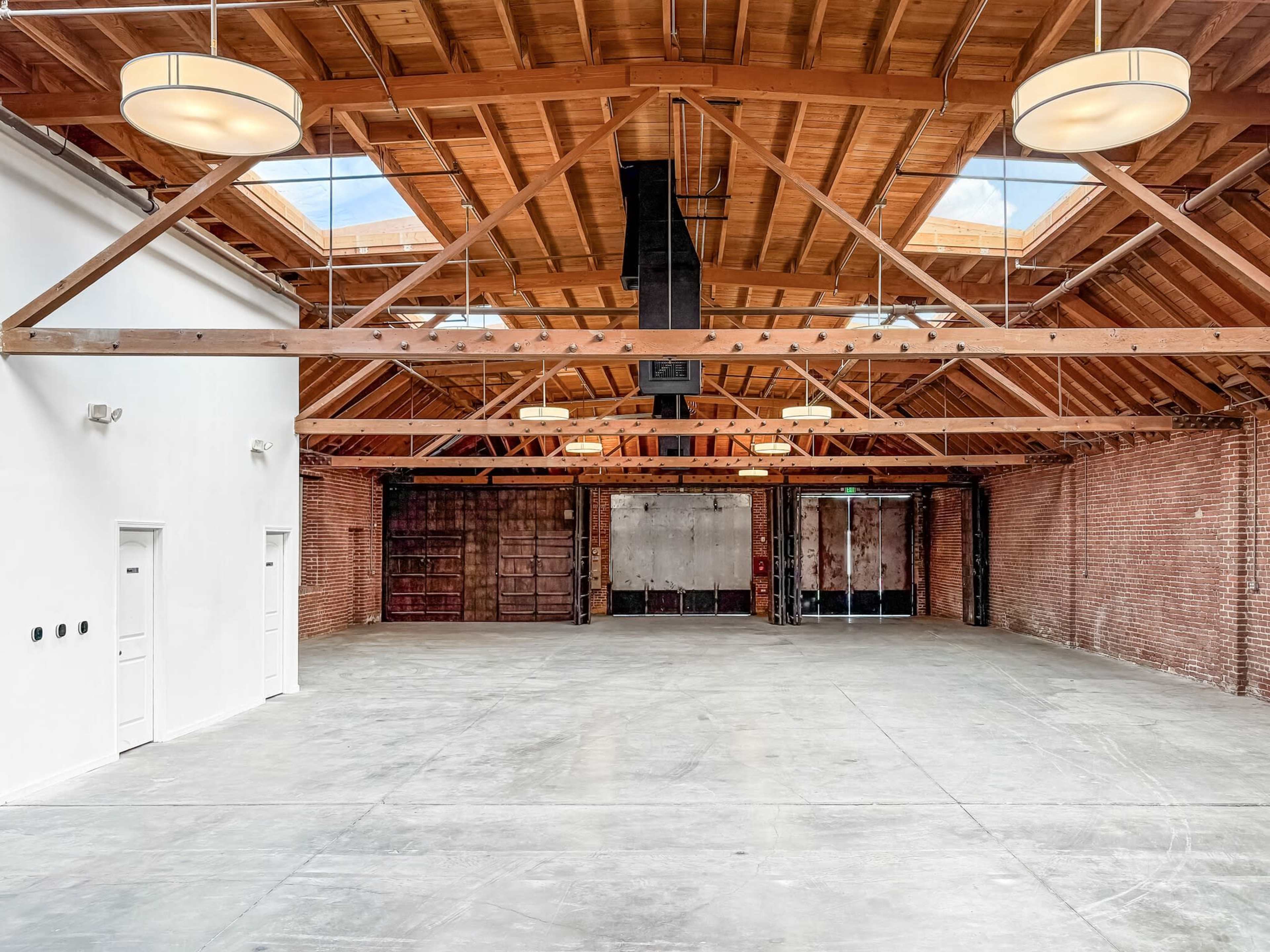 The image shows an empty industrial space with exposed brick walls and a wooden ceiling, featuring large skylights and concrete flooring.