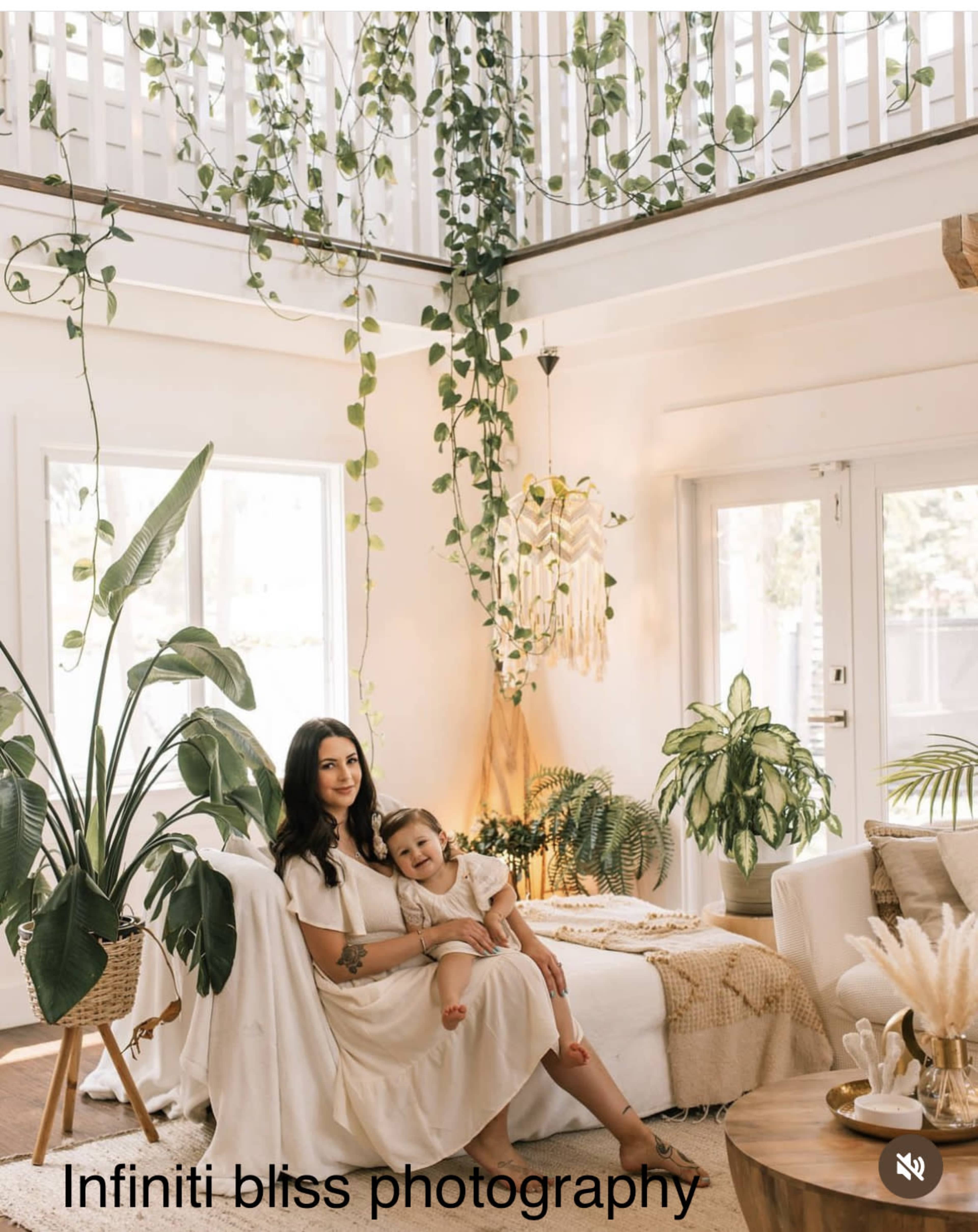 A woman sits on a white sofa in a brightly lit room adorned with hanging plants and decorative elements, holding a young child in her lap.