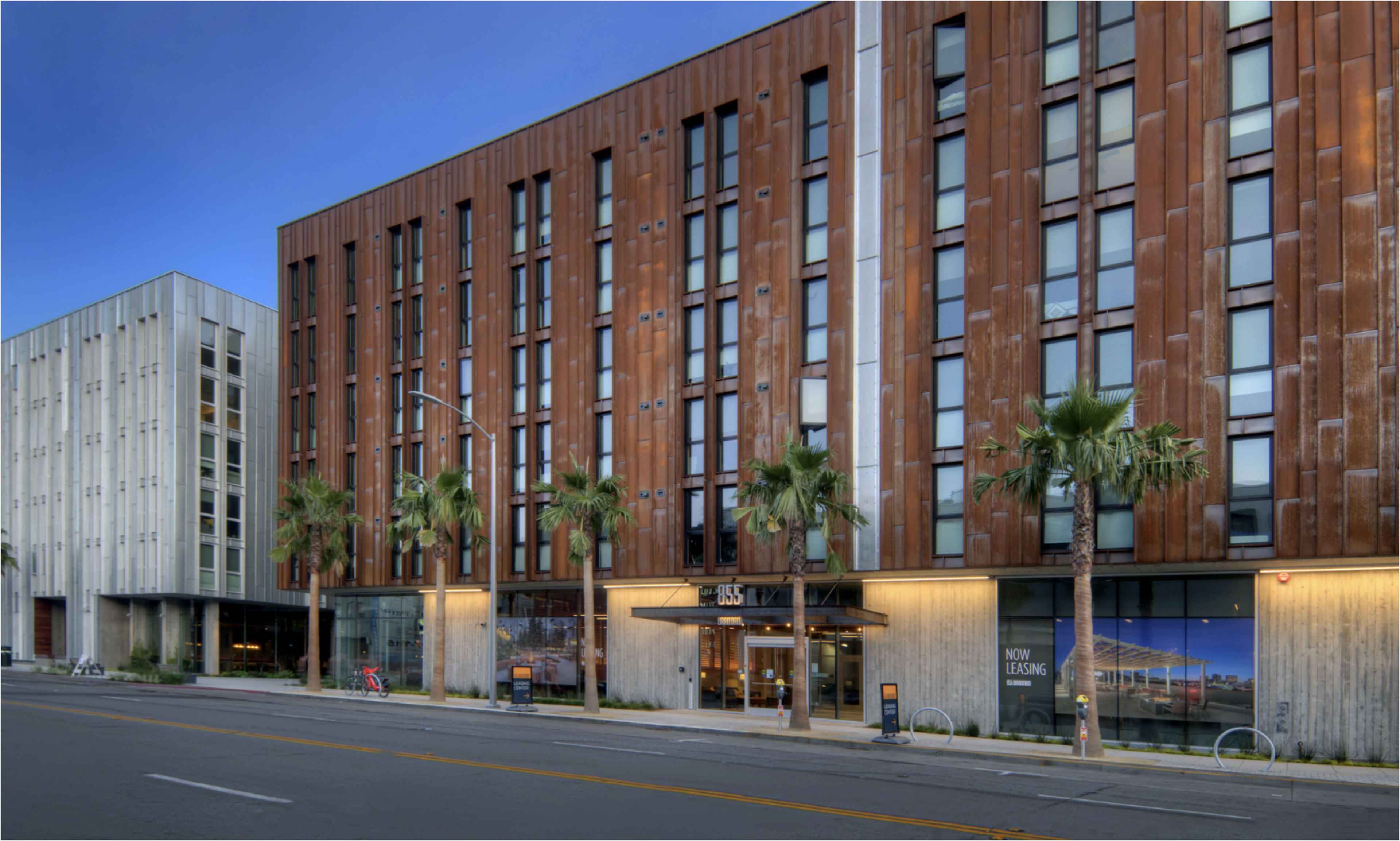 The image shows two modern multi-story buildings, one with a rust-colored facade and large windows, and the other with a lighter exterior, both situated along a street lined with palm trees.