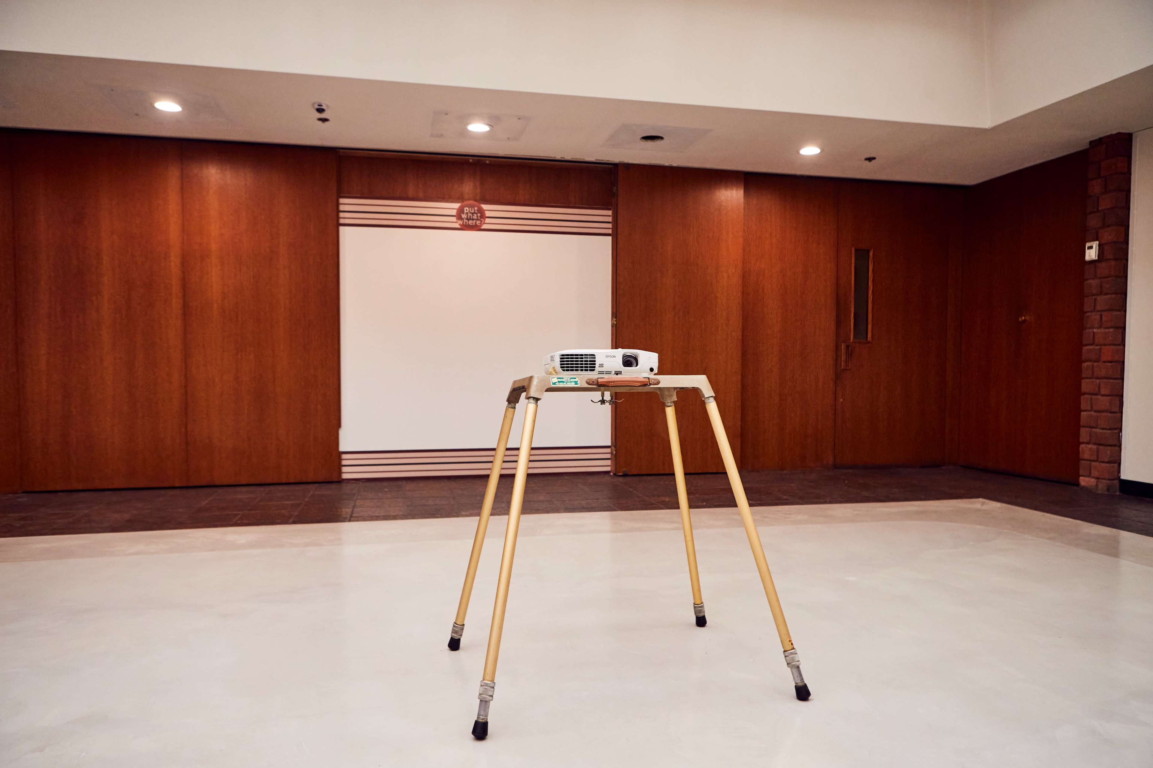 A projector is mounted on a tripod in an empty room with wooden paneling and a blank screen.