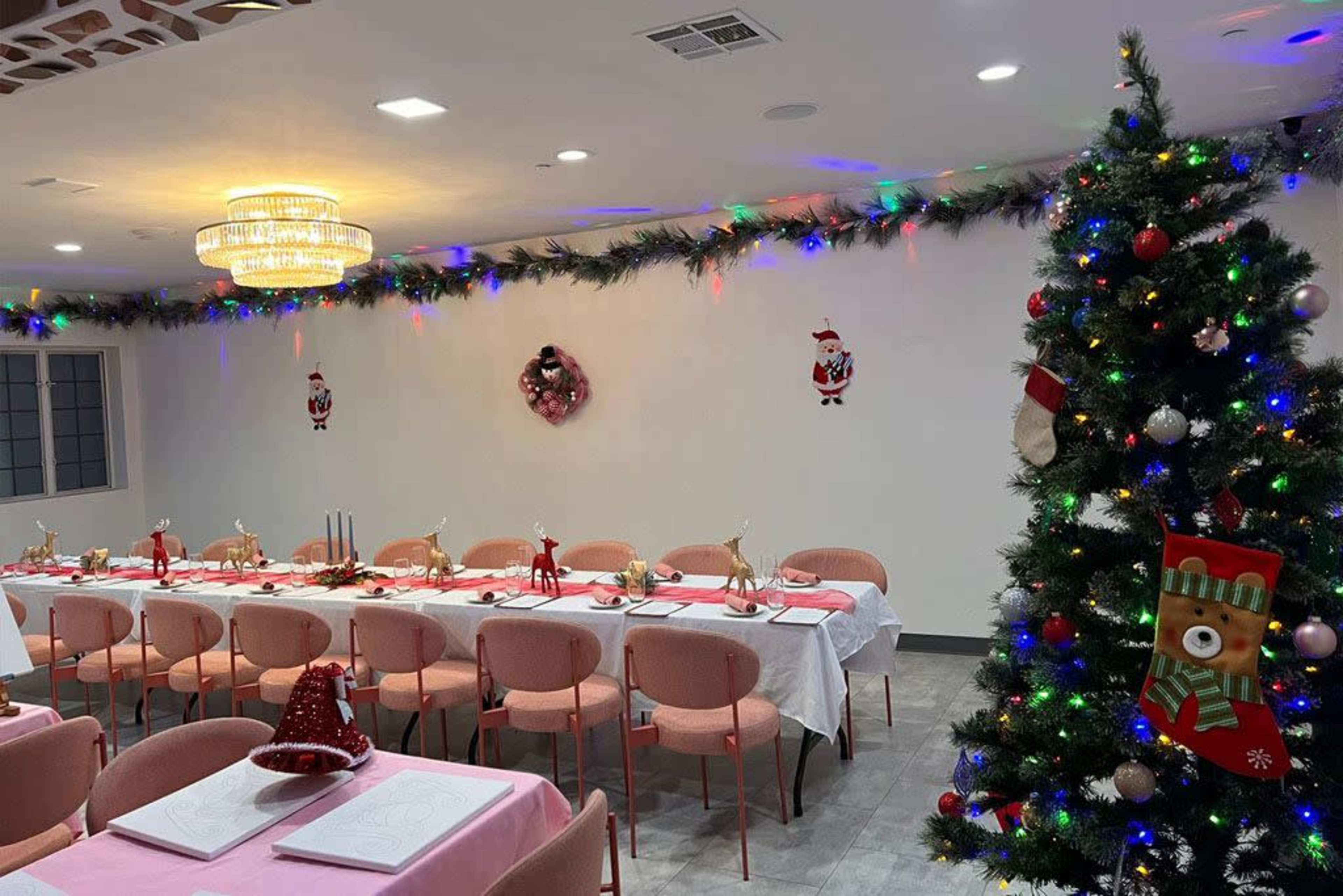 A festive dining room is set up for a holiday gathering, featuring a long table adorned with pink tablecloths and decorations, surrounded by pink chairs, with a Christmas tree in one corner and holiday decorations on the walls.