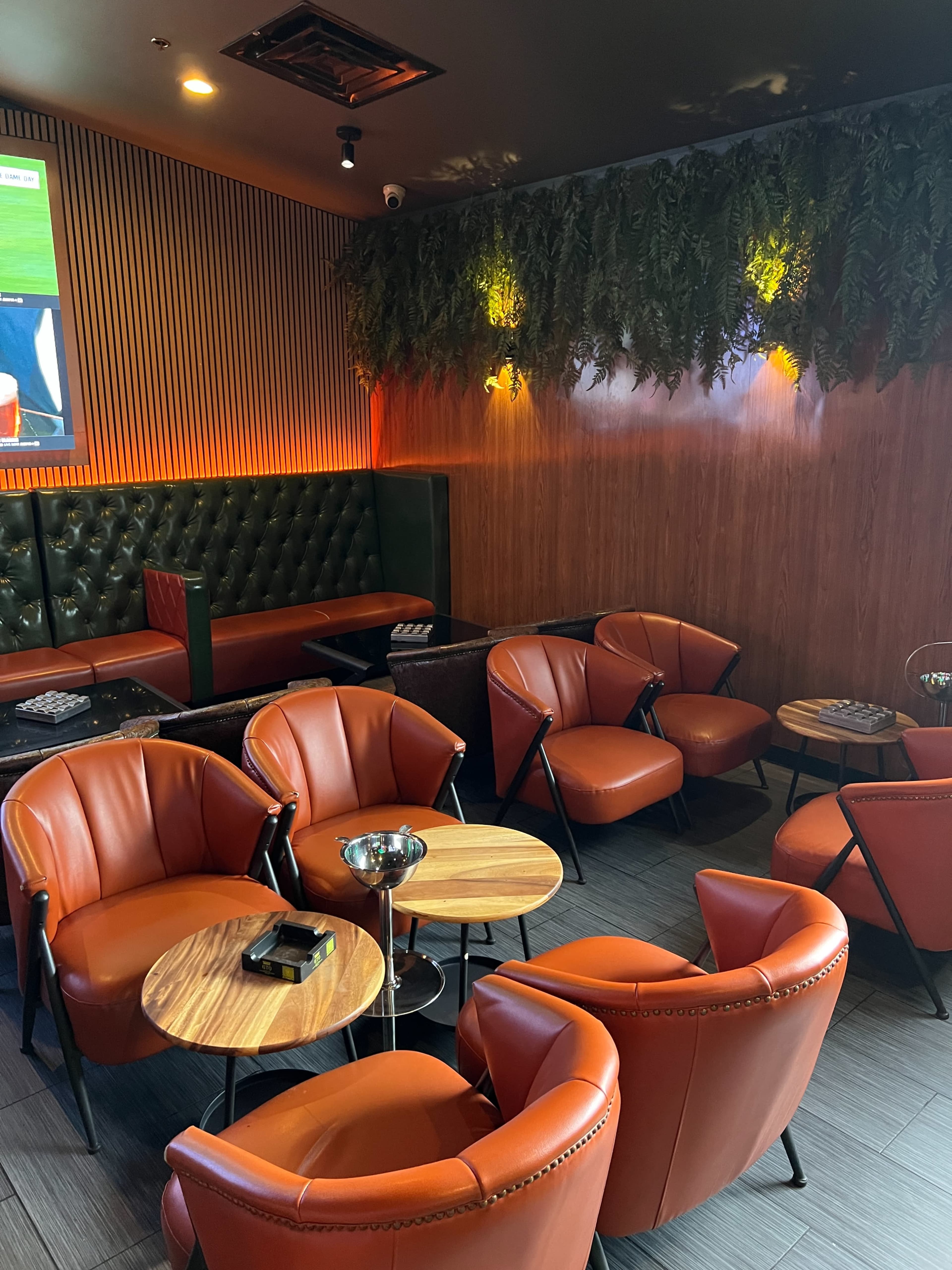 The image shows a cozy lounge area with orange leather chairs, wooden tables, and textured walls, featuring a large screen in the background.