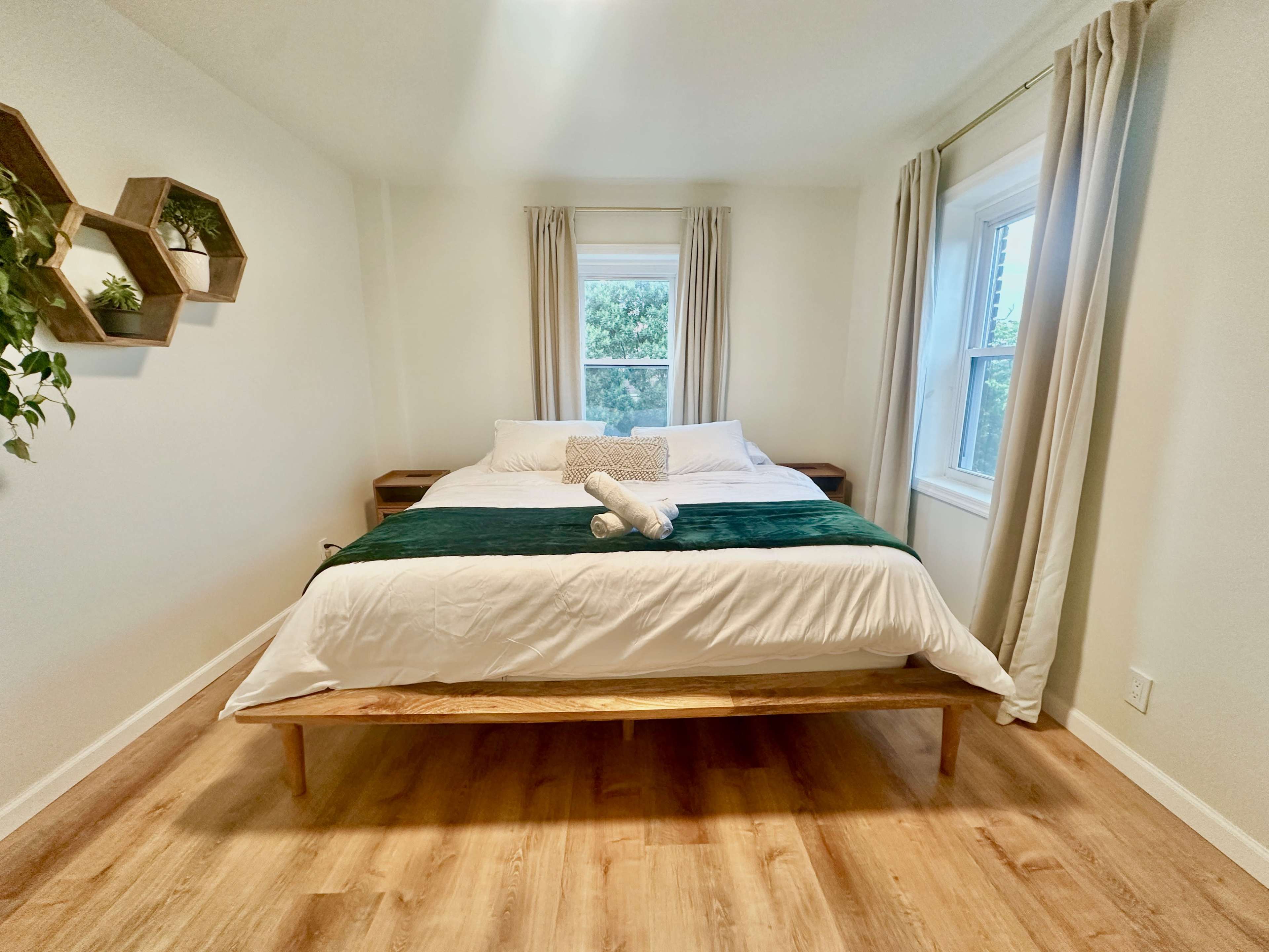 A neatly arranged bedroom features a large bed with white bedding and green accents, positioned between two windows, and complemented by a wooden bed frame and wall decor.