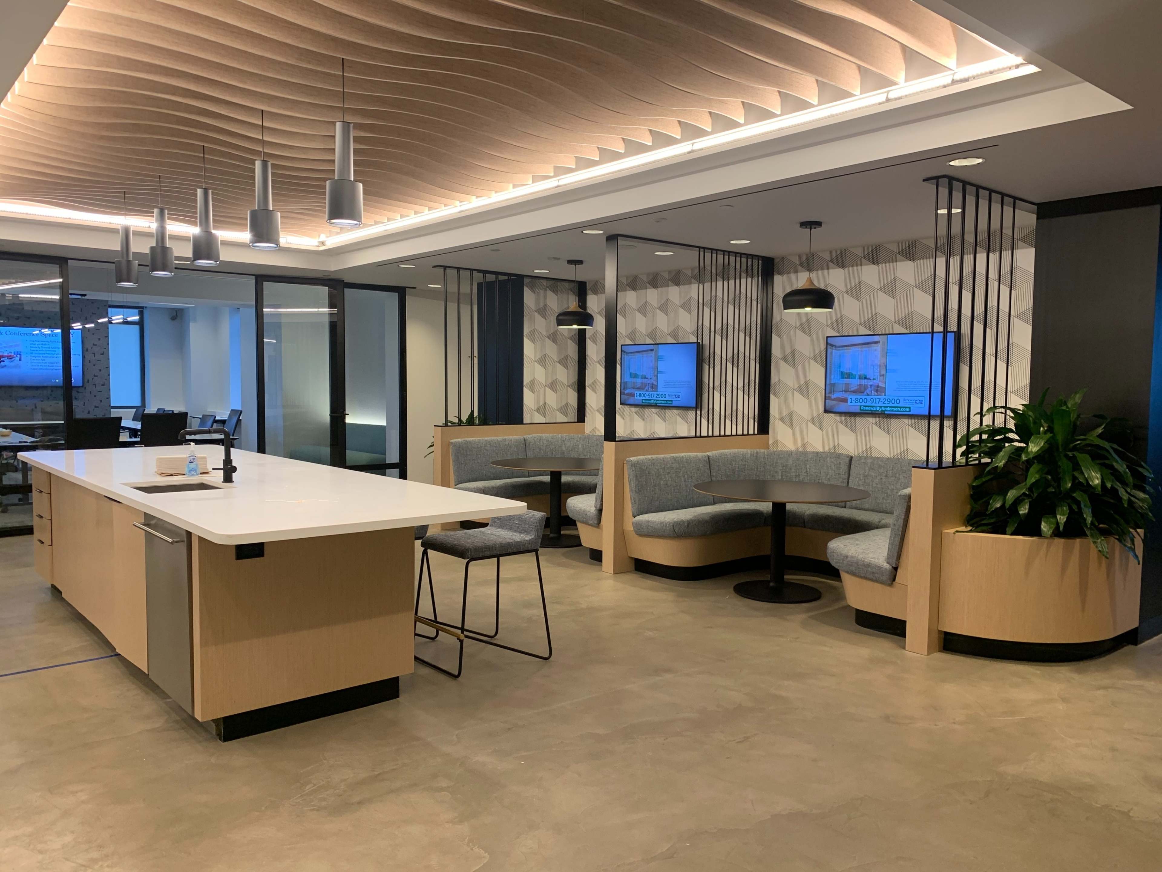 The image shows a modern office interior with a seating area featuring curved booths, a white countertop island, and two wall-mounted televisions.